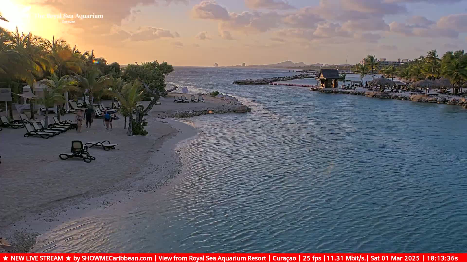 A tranquil beach scene at sunset shows a sandy shoreline with lounge chairs and people walking, alongside calm teal water leading to a distant coastline under a partly cloudy sky.