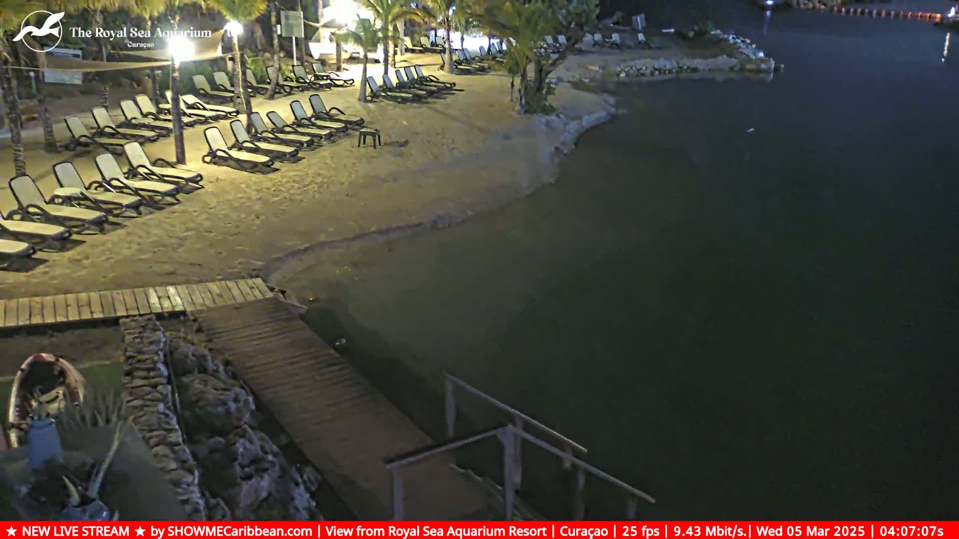 Empty lounge chairs sit on a sandy beach at night next to a calm body of dark water, with a wooden walkway leading to the water.