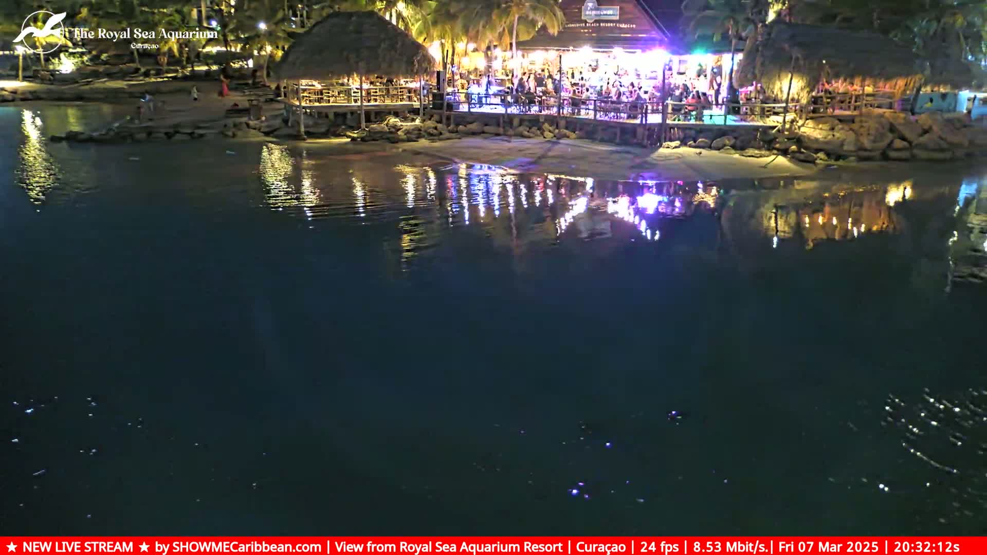 A brightly lit waterfront restaurant at night is reflected in calm, dark water.