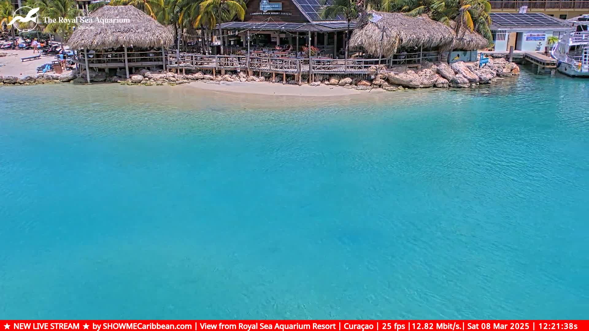 A beachside restaurant with thatched roofs sits on a sandy shore next to calm, bright turquoise water.