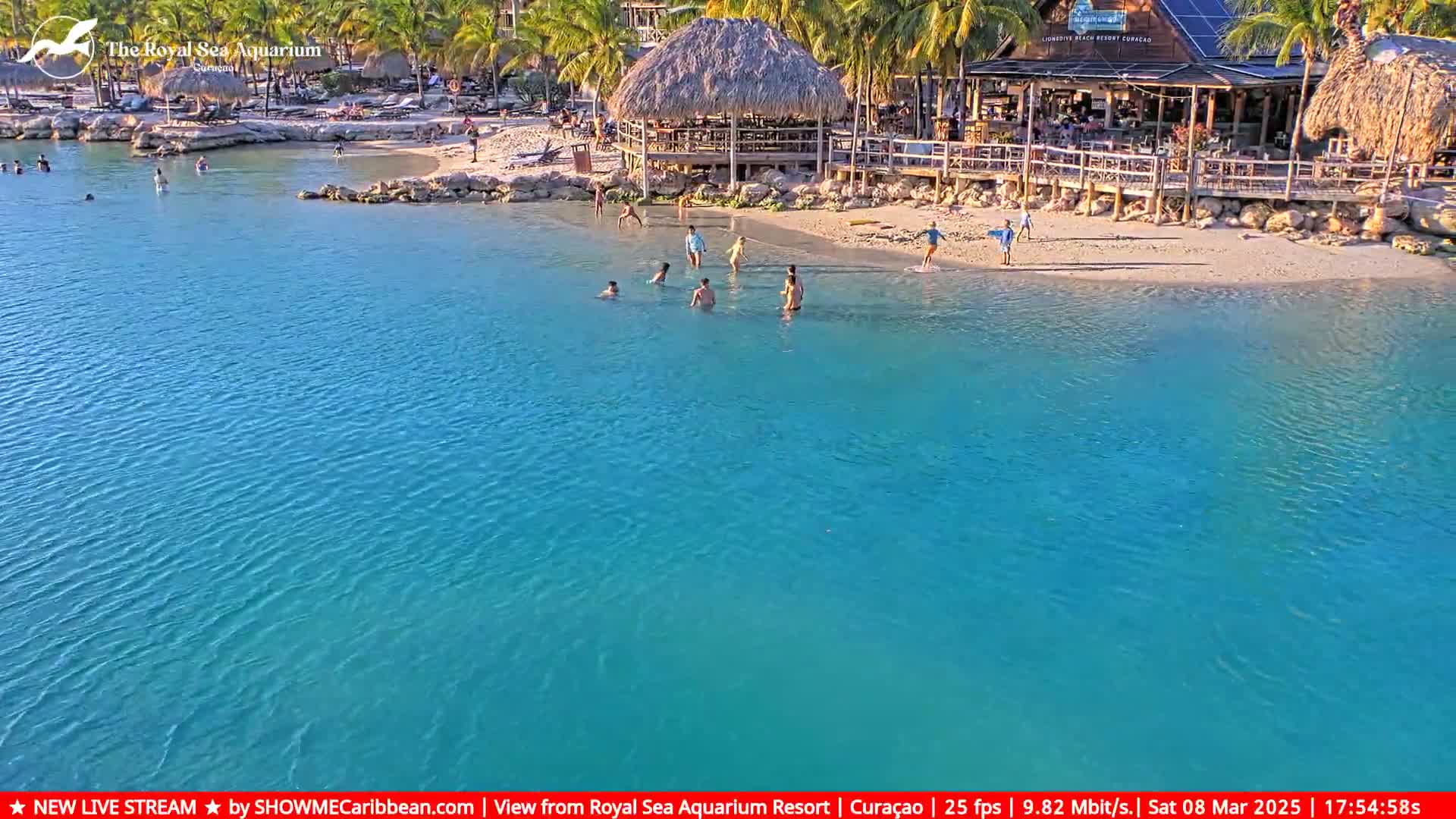 A calm, turquoise body of water laps a sandy beach where several people are swimming and wading near a beachfront restaurant and bar under a sunny sky.