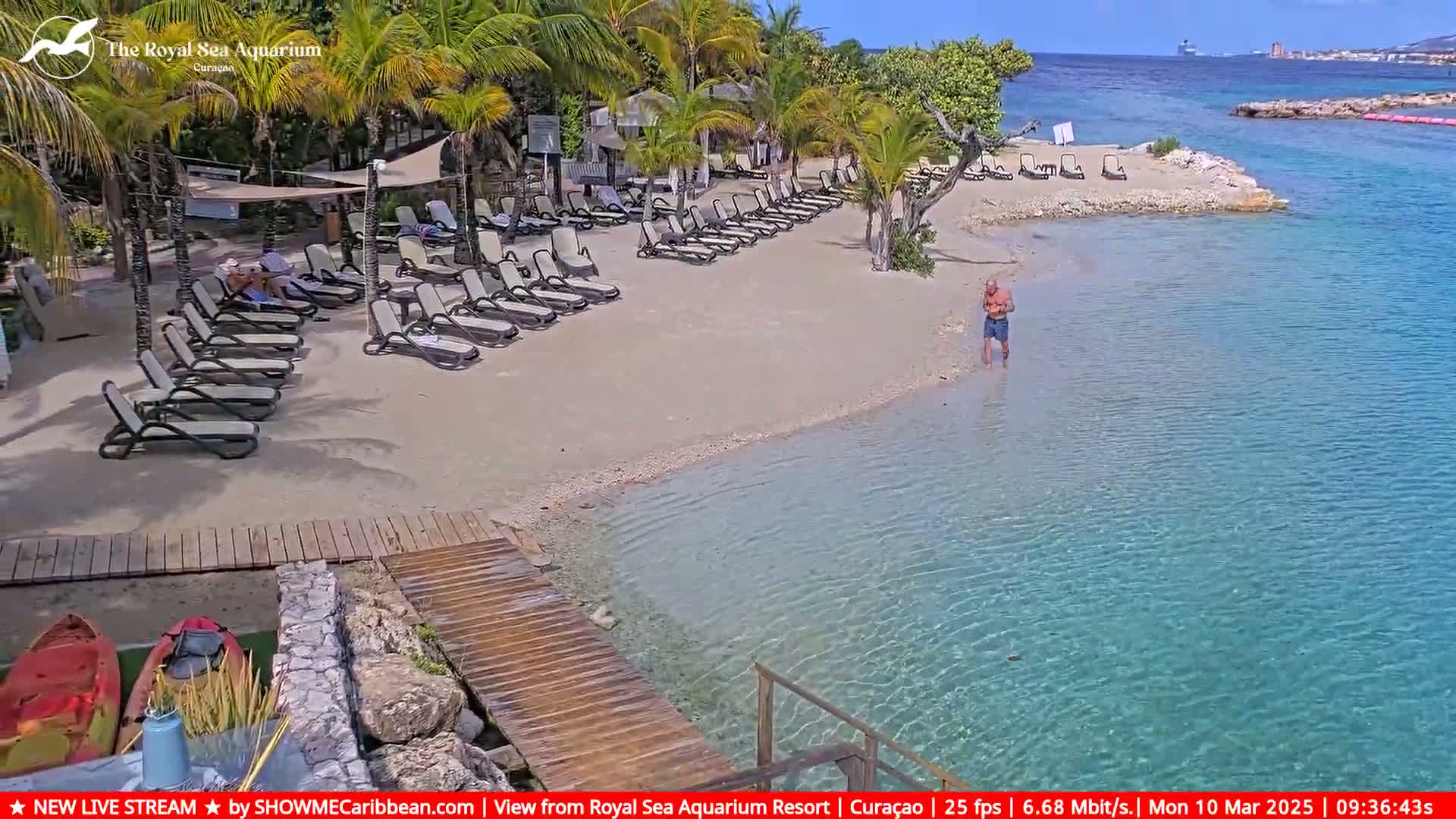 A sandy beach with lounge chairs and a man wading in clear, shallow turquoise water under a sunny sky.
