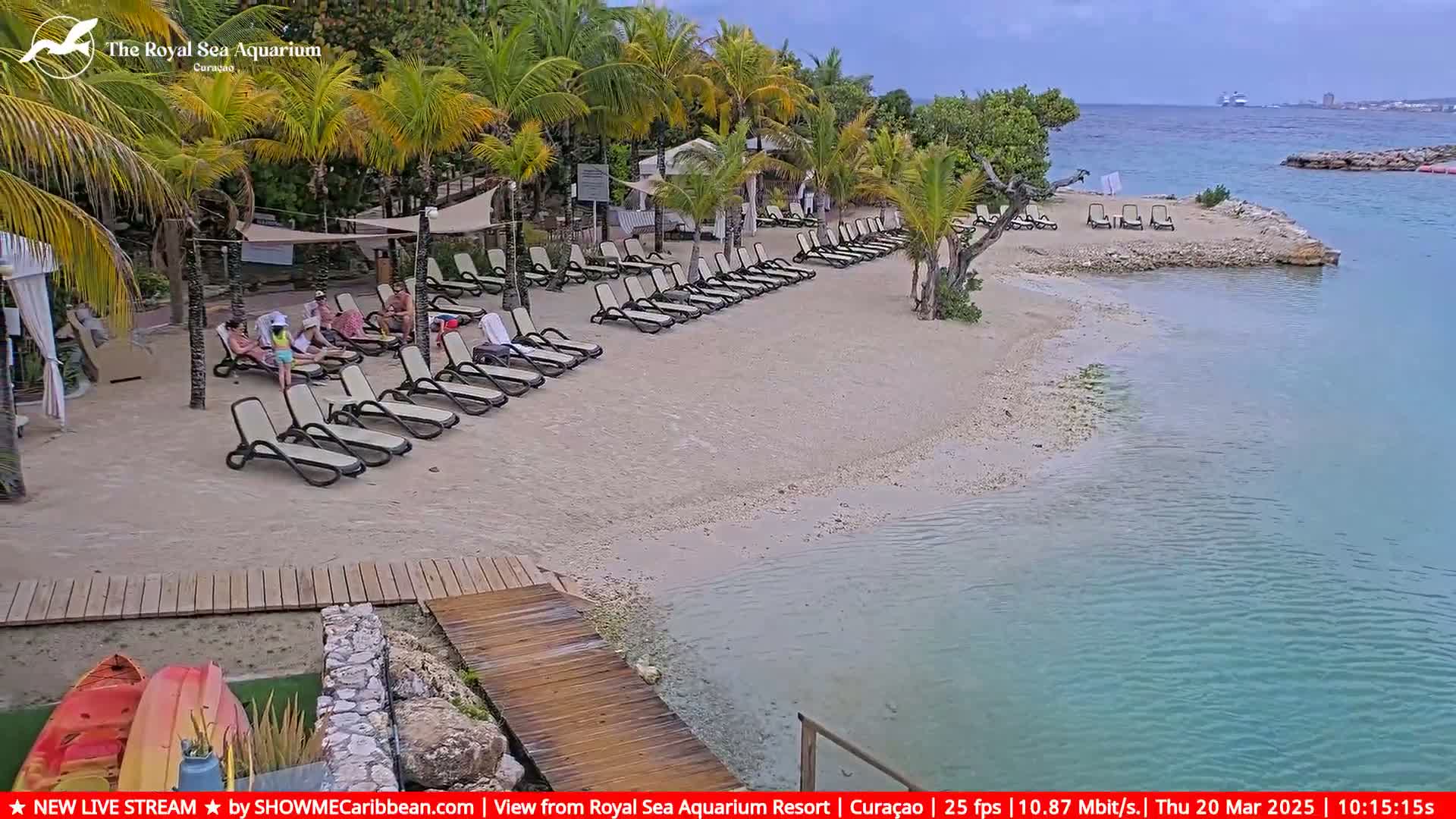A sandy beach with lounge chairs and several people under cloudy skies, next to calm turquoise water and palm trees.