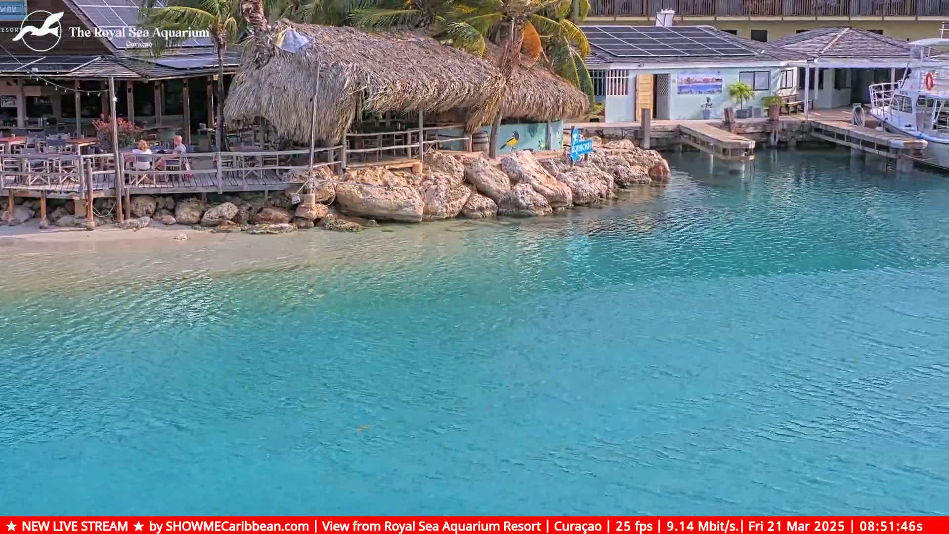 A waterfront restaurant with thatched roof sits on a rocky shore next to calm, turquoise water.