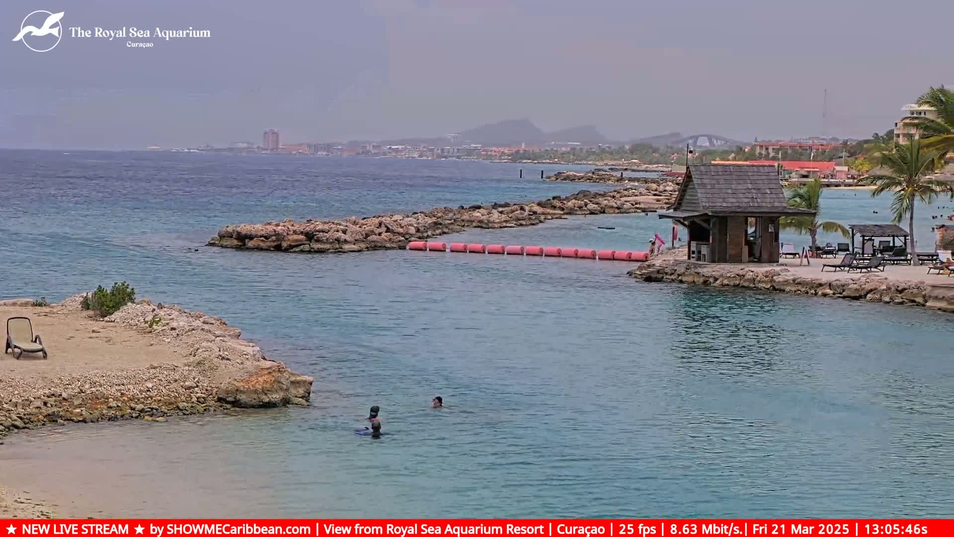A hazy, sunny day at a beachside resort shows a calm body of water with rocky inlets, a small wooden hut, lounge chairs, and a few people swimming.
