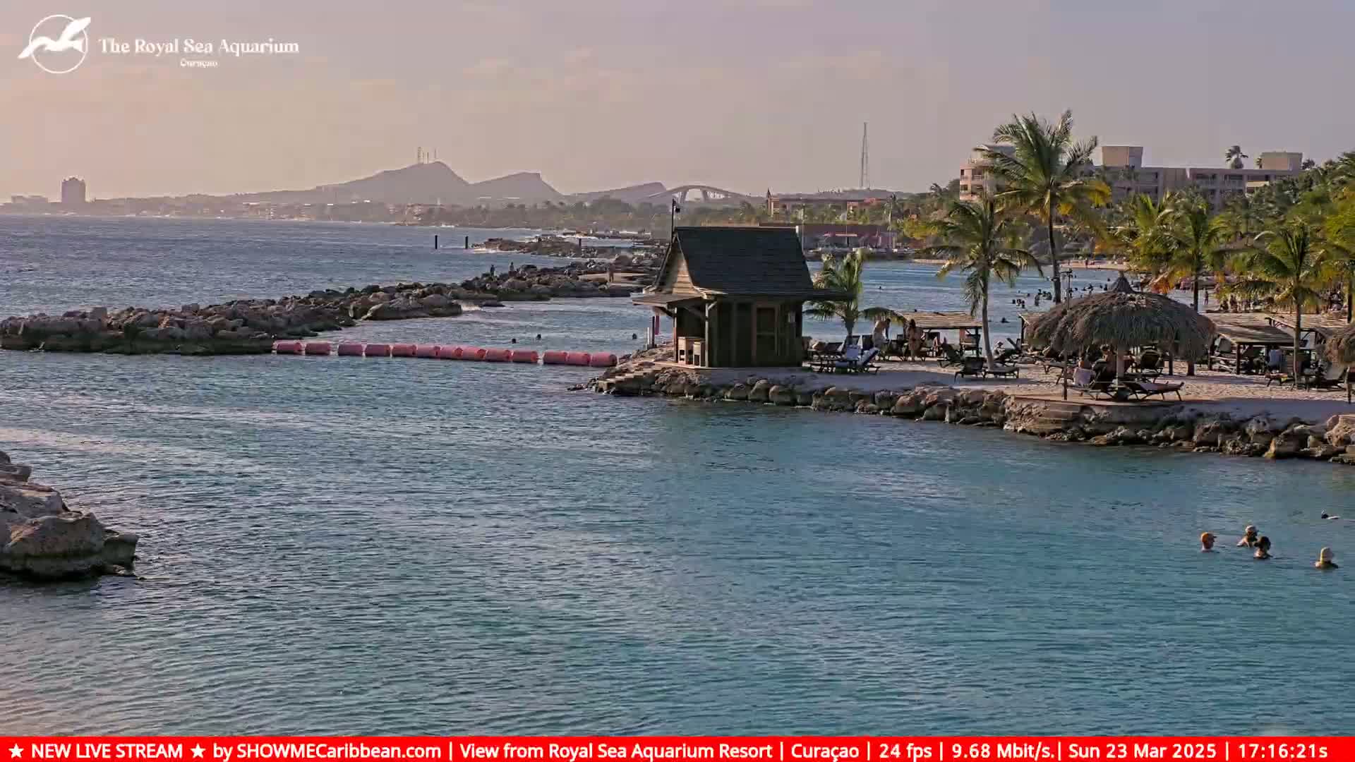 A calm, sunny day at a beach resort shows a small wooden structure on a rocky peninsula, with people swimming and relaxing on a sandy beach under palm trees and thatched umbrellas, and a distant view of hills and a bridge.