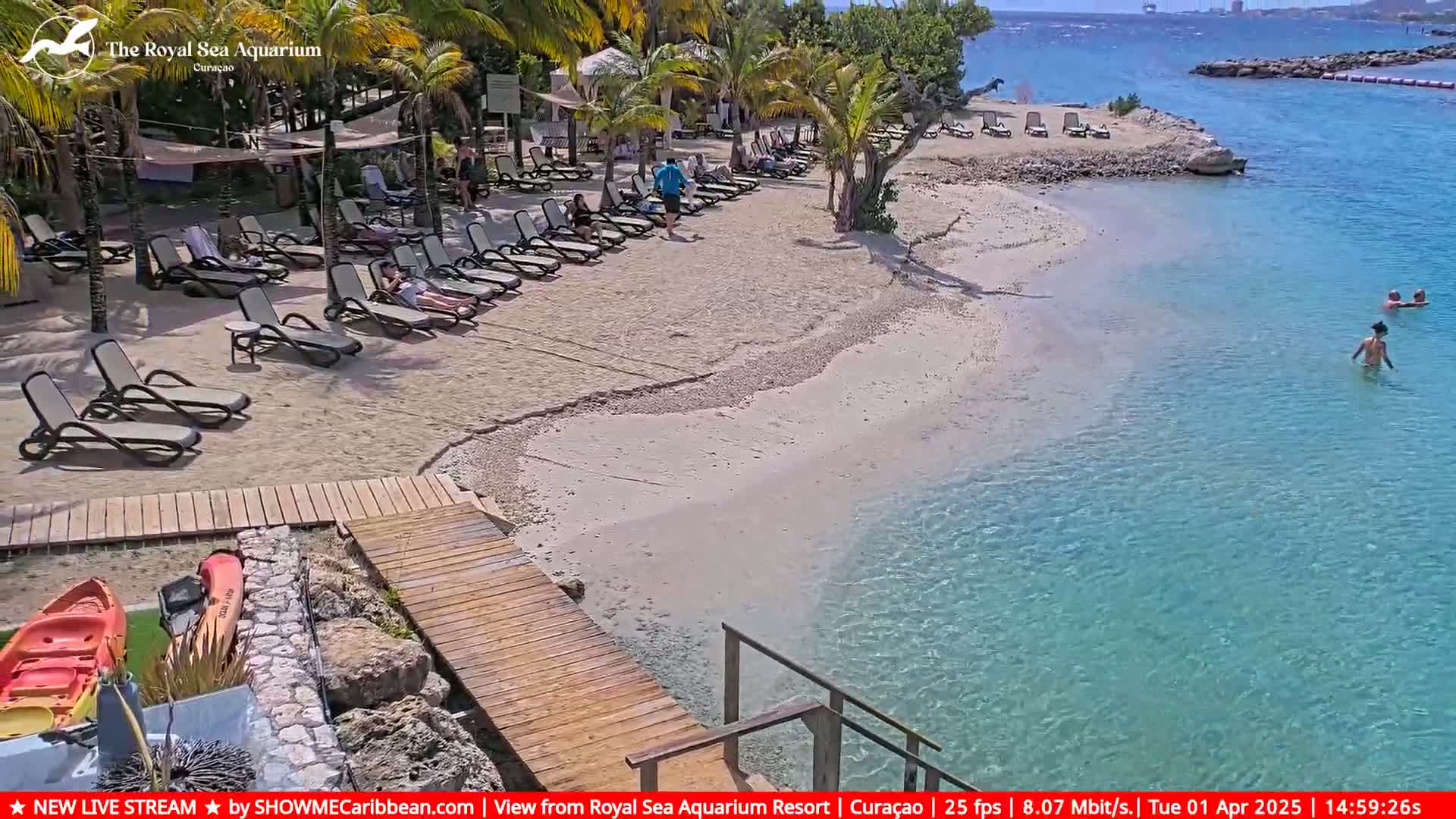 A sandy beach with lounge chairs and a few people, leading to clear turquoise water under a sunny sky.