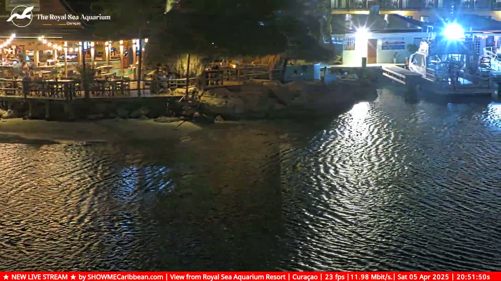 A nighttime scene shows a waterfront restaurant with outdoor seating and string lights, next to a calm body of water with a boat docked nearby.