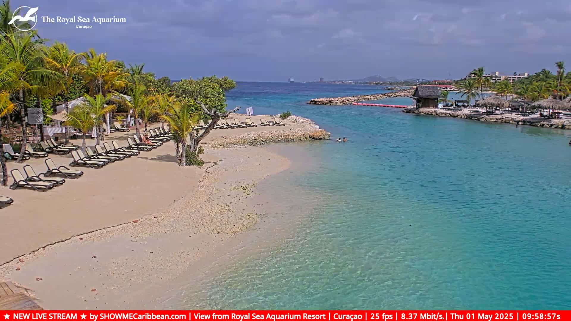 A sandy beach with lounge chairs is next to a calm, clear turquoise body of water under a partly cloudy sky.