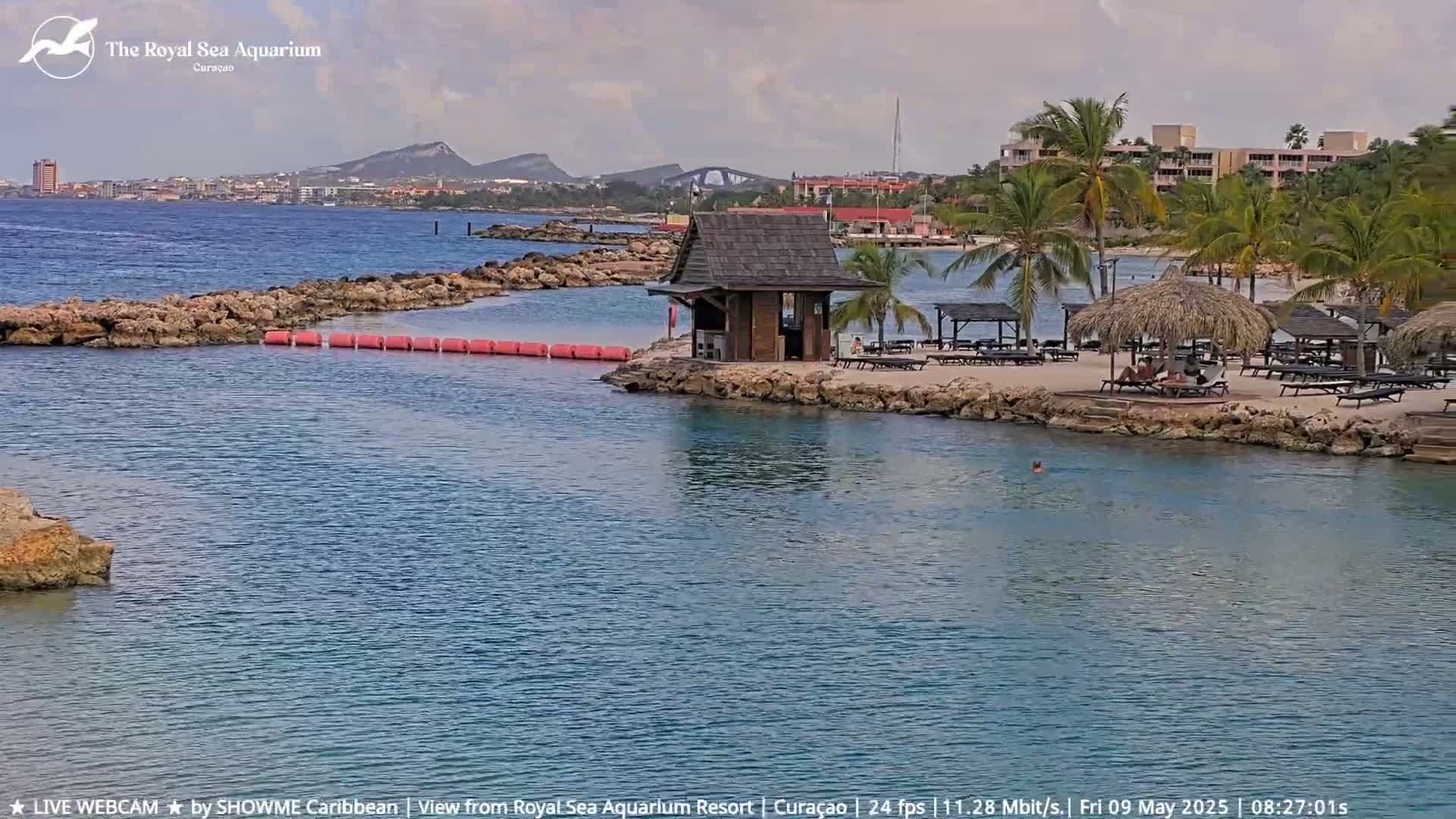 A calm, partly cloudy day reveals a beach scene with a small wooden hut, palm trees, lounge chairs, and a rocky shoreline bordering turquoise water.
