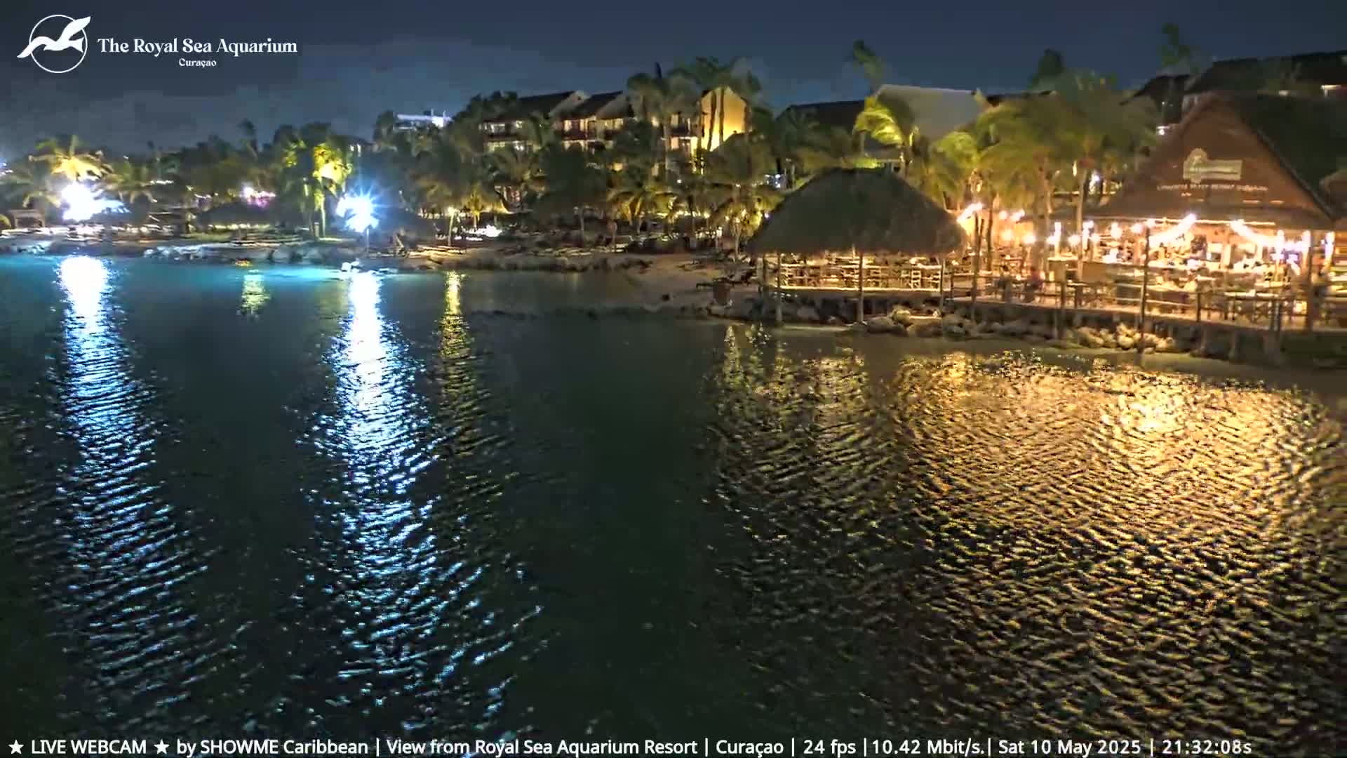 A nighttime view of a calm body of water reflecting lights from a resort with palm trees and a beachfront restaurant.