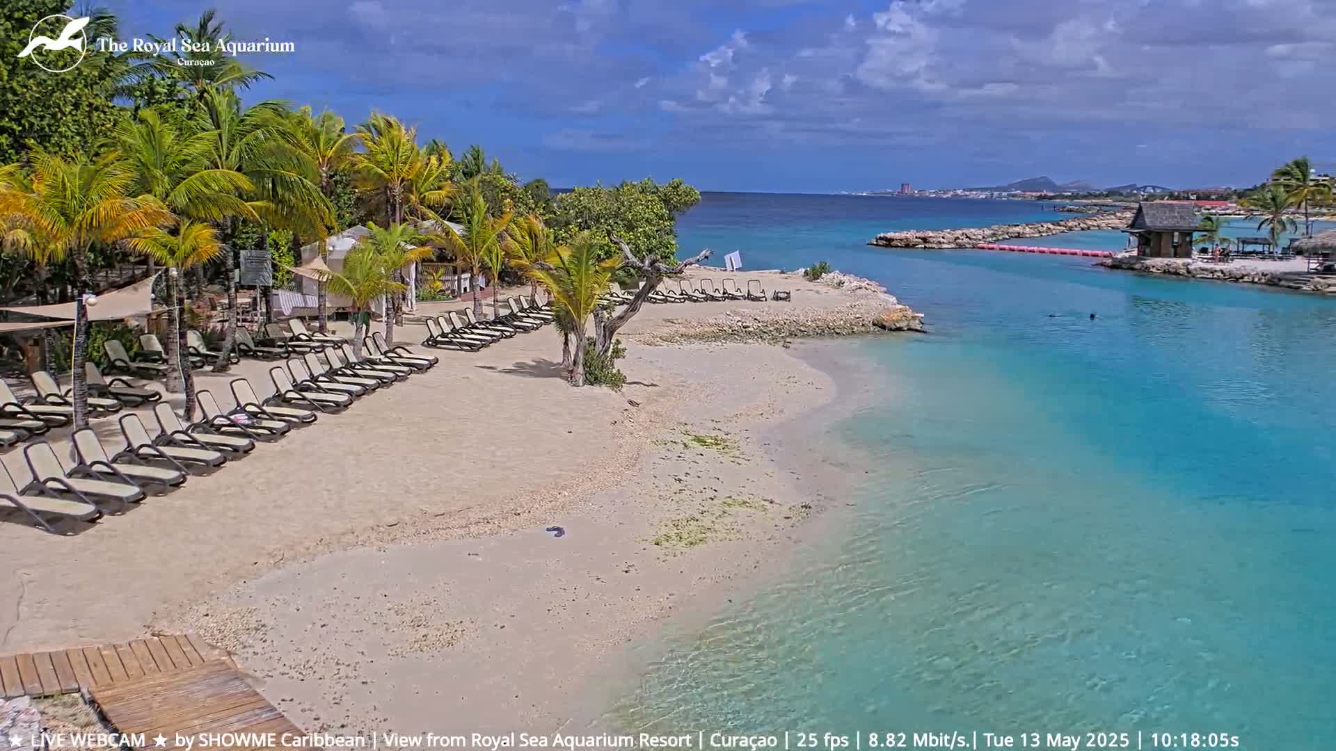 A sandy beach with lounge chairs is nestled beside a calm, turquoise body of water under a partly cloudy sky.