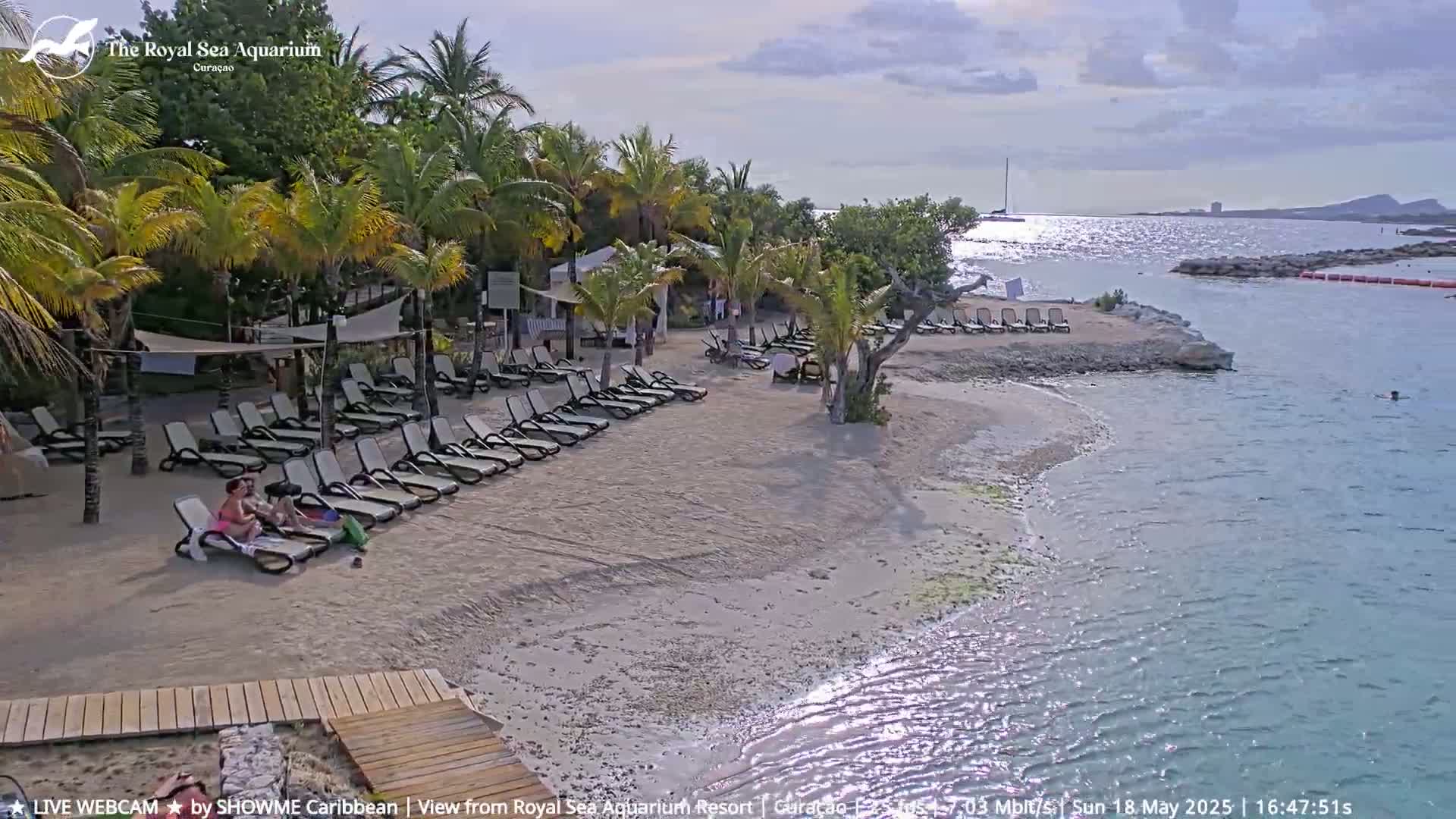 A sandy beach with lounge chairs, palm trees, and a few people is partially surrounded by calm, sparkling water under a partly cloudy sky.