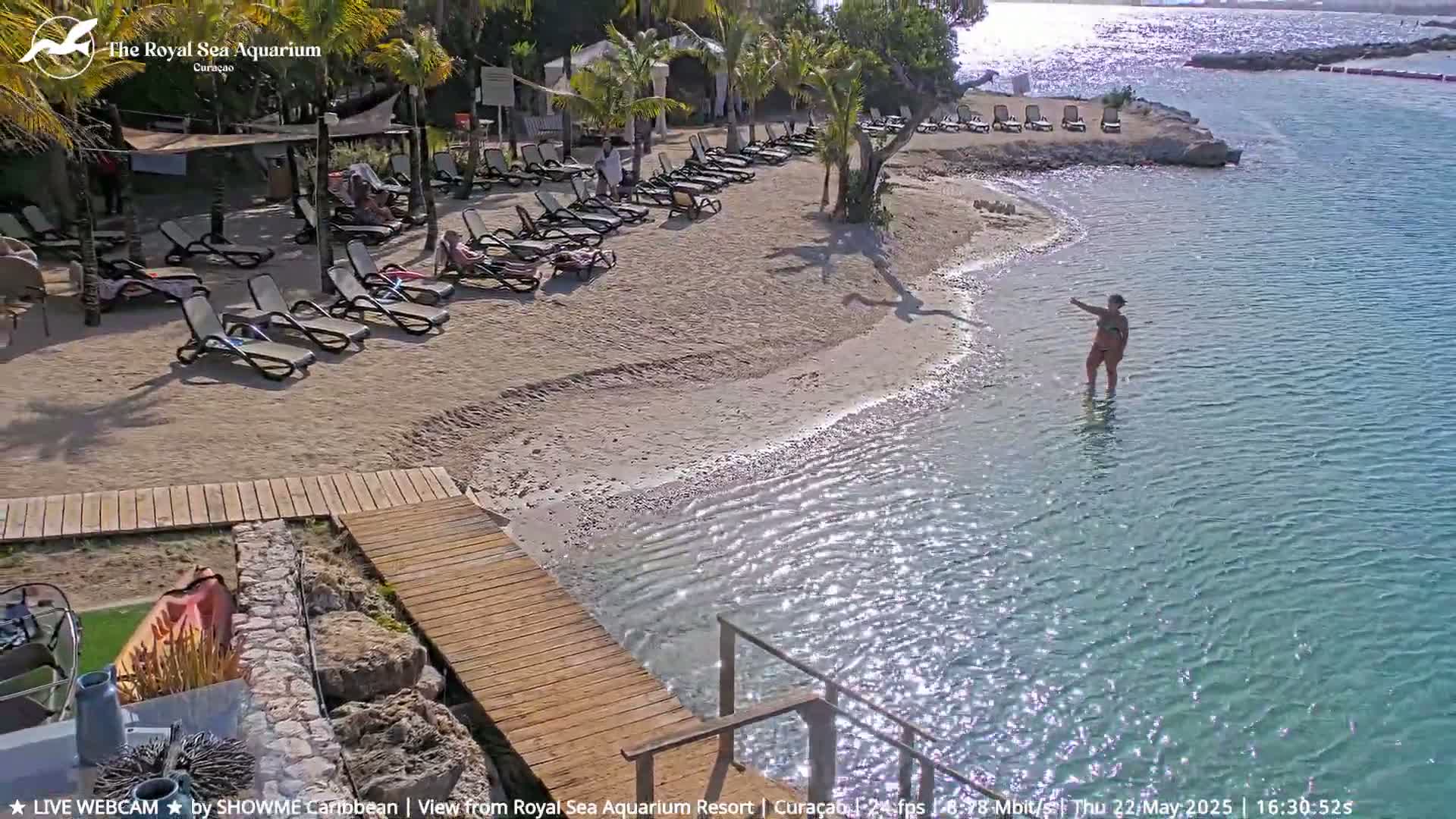 A sandy beach with lounge chairs and a woman wading in calm, clear turquoise water under a sunny sky.