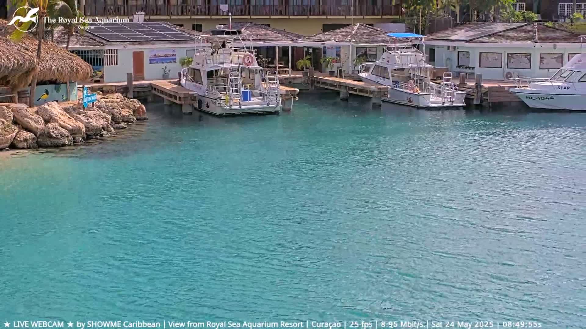 Two boats are docked at a marina with calm, teal water, next to a building with a thatched roof.