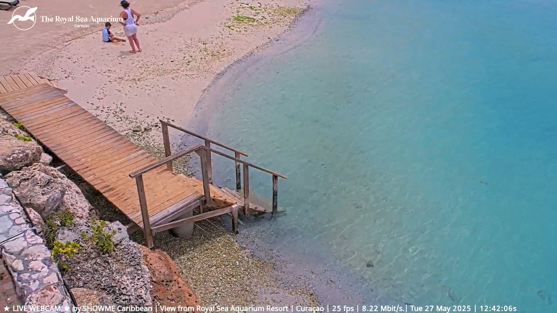 A wooden dock with stairs leads down to clear, shallow turquoise water from a sandy beach where a woman and child are playing.