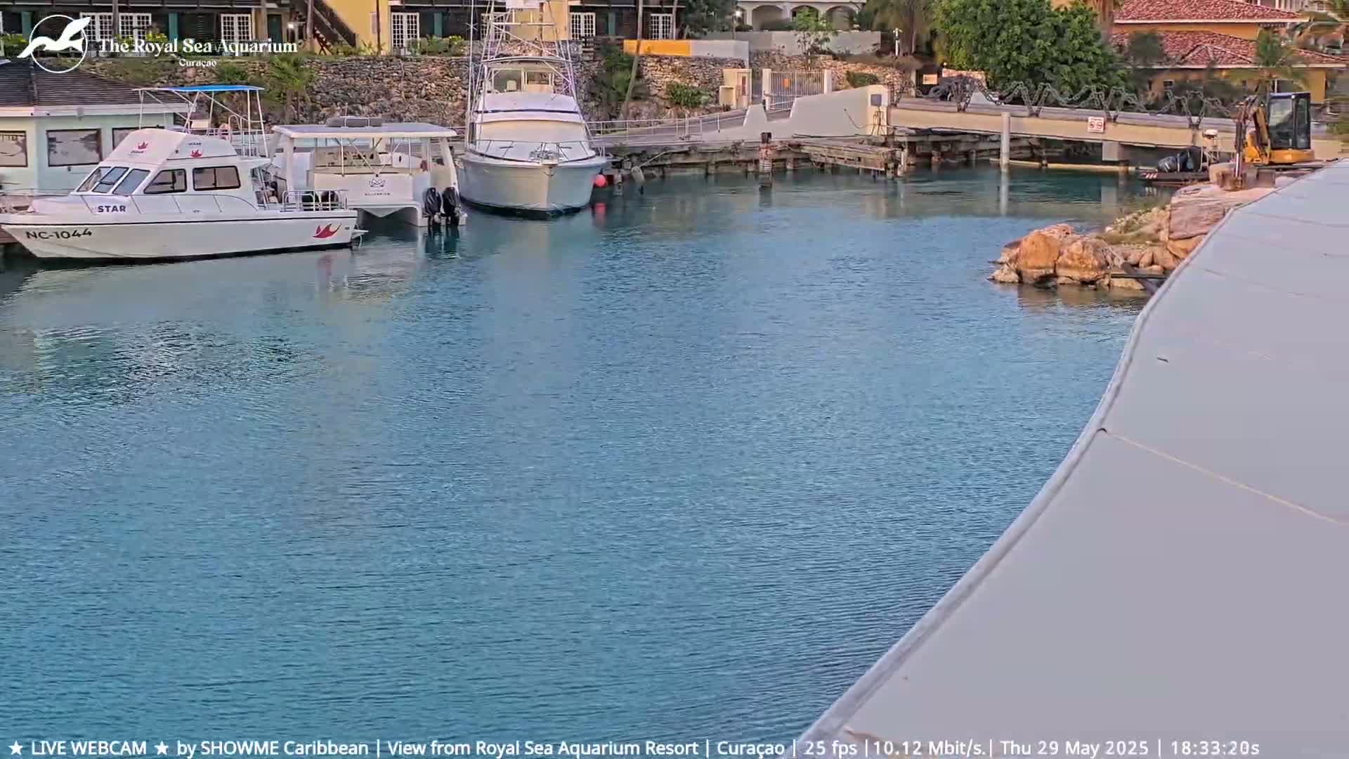 Several boats are moored in a calm, blue harbor next to a rocky shoreline and a small, yellow excavator.