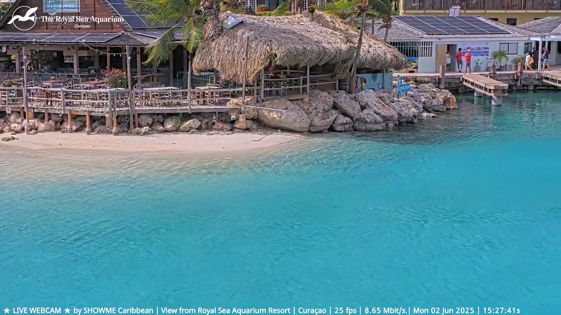 A beachfront restaurant with a thatched roof sits on a sandy shore next to clear, turquoise water.