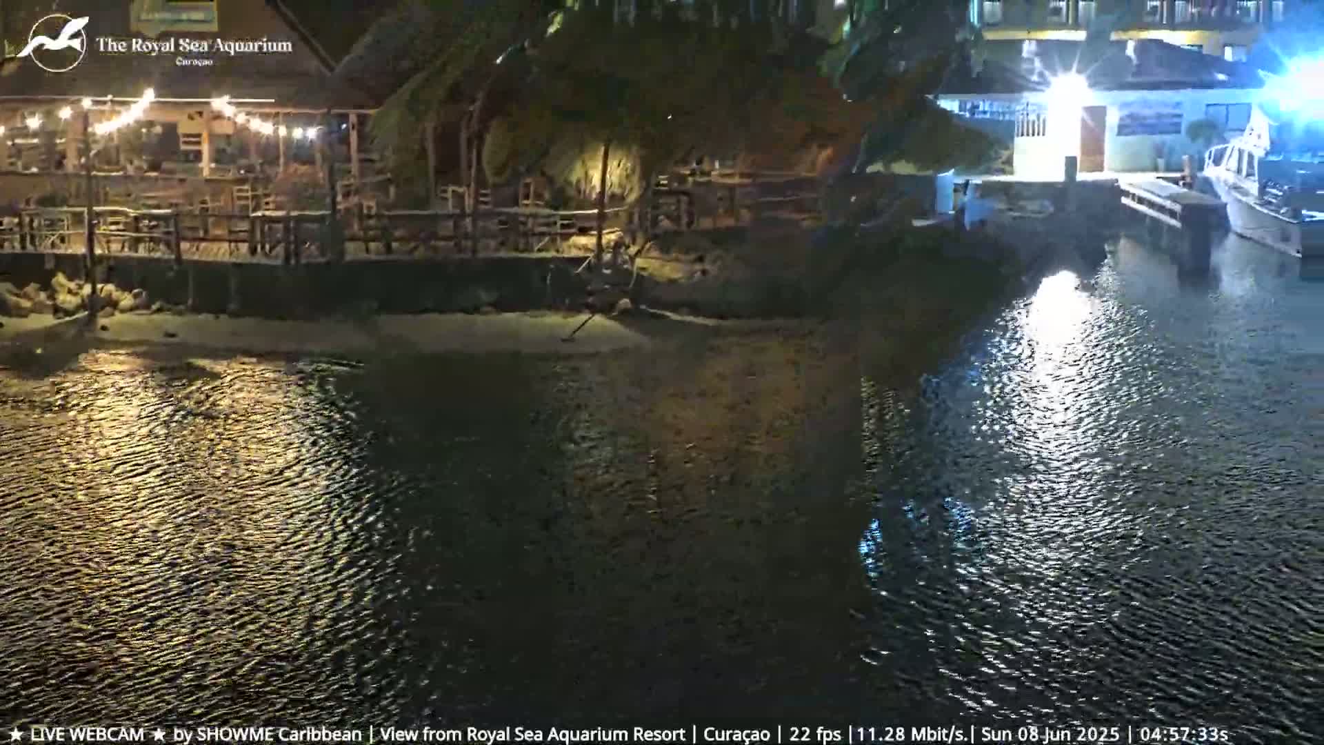 A nighttime view of a waterfront restaurant with string lights, a sandy beach, and a calm body of water reflecting lights from nearby buildings and boats.