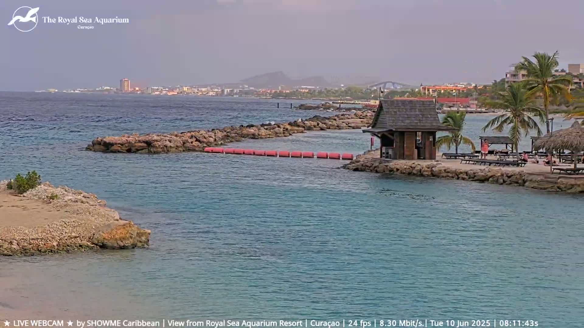 A calm, partly cloudy day at a beach resort shows a small cove with a wooden structure and lounge chairs, a rocky barrier, and a distant cityscape under a hazy sky.