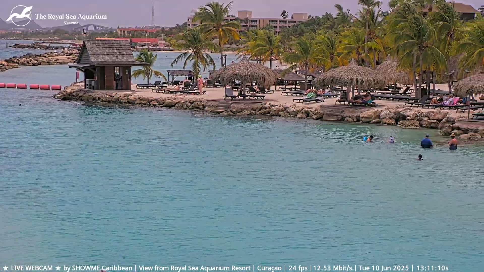 A tranquil beach scene shows a sandy shore with thatch-roofed cabanas and people relaxing, a small wooden building, and calm, shallow turquoise water with a few swimmers.