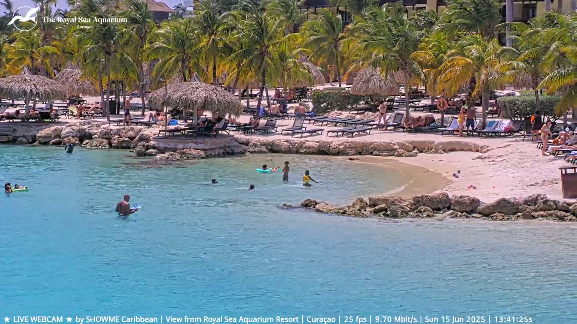 A sandy beach with palm trees, thatched umbrellas, and lounge chairs surrounds a calm, turquoise body of water where several people are swimming and sunbathing under a sunny sky.