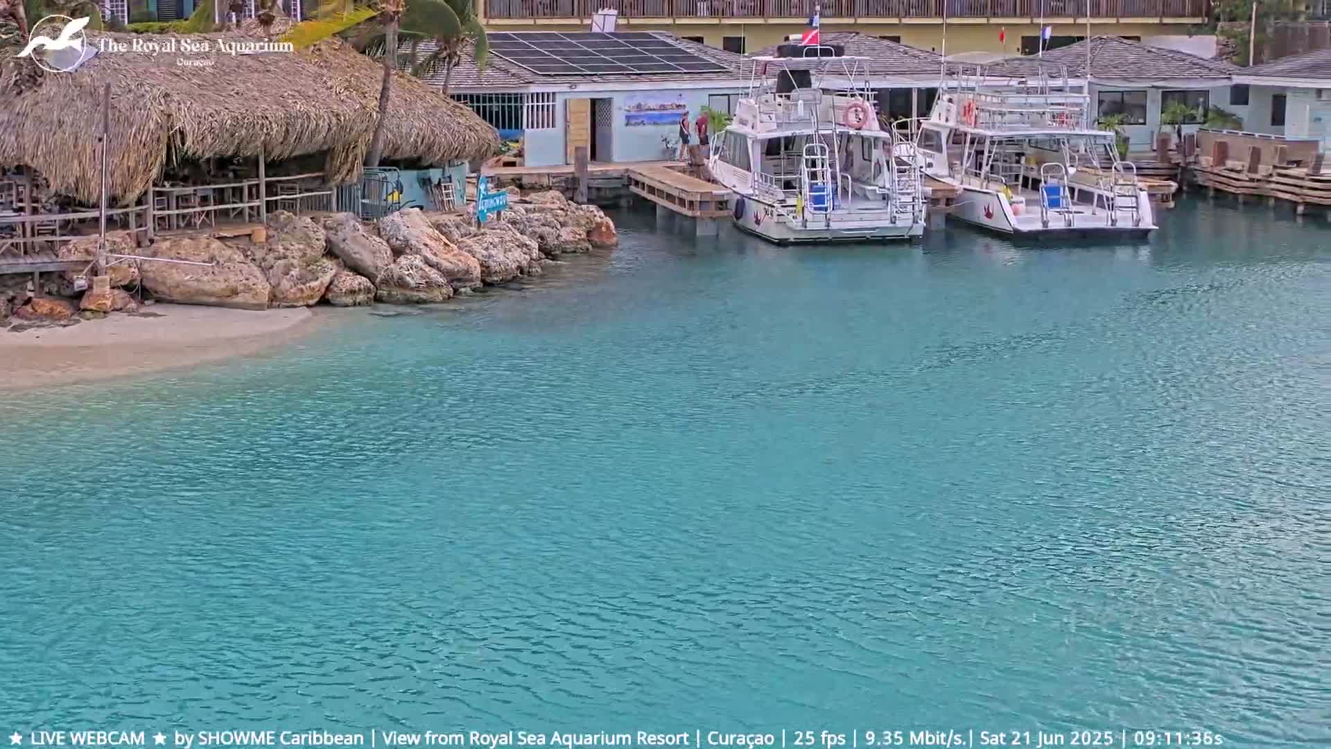 Two boats are docked at a waterfront building near a thatched-roof structure, with calm, clear turquoise water surrounding them.