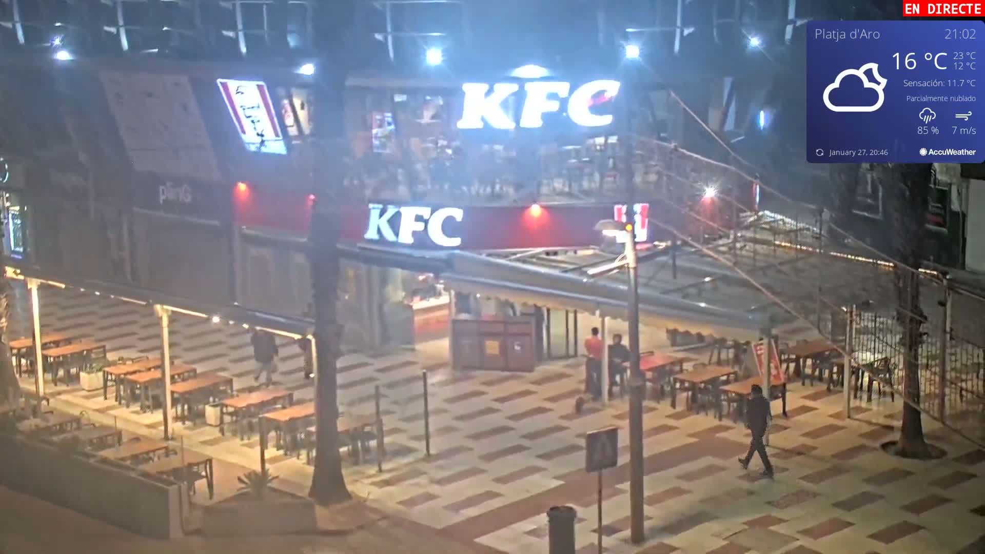 A nighttime, partly cloudy scene shows a KFC restaurant with outdoor seating, a few people walking around, and a lightly hazy atmosphere.