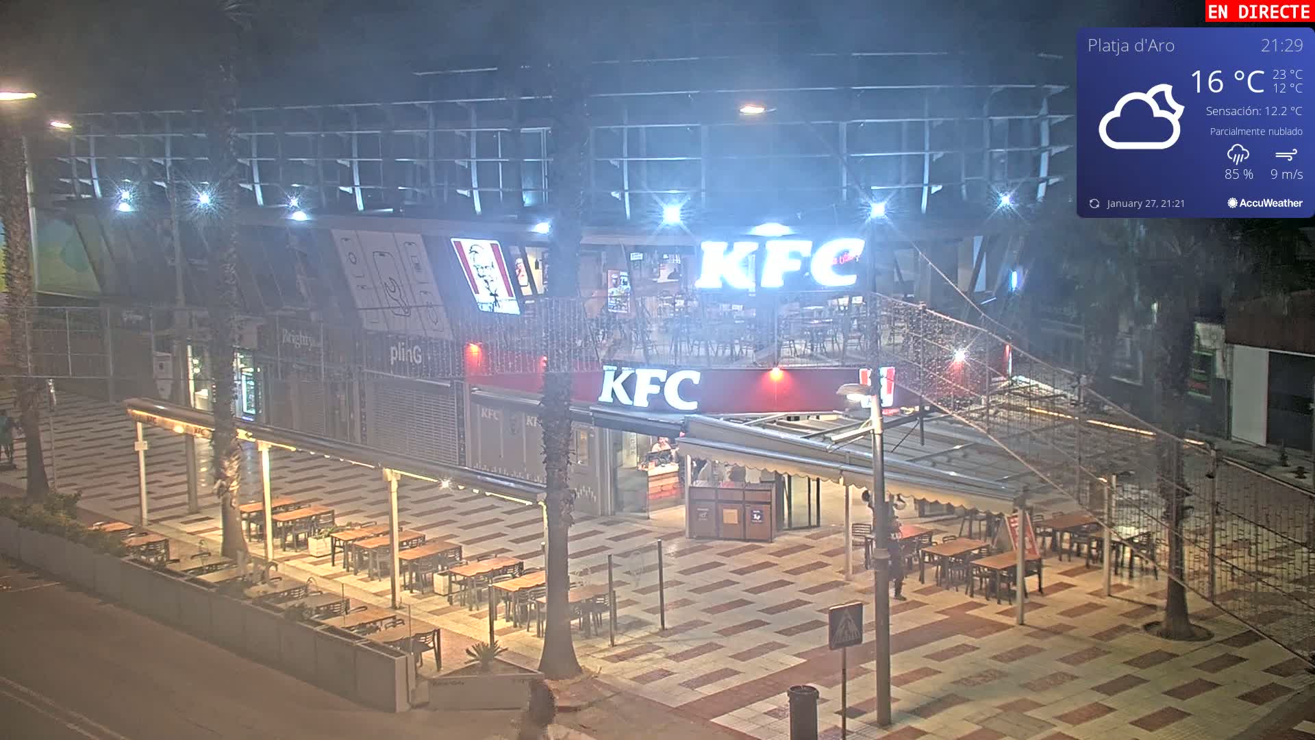 A nighttime view of an outdoor seating area in front of a KFC restaurant under partly cloudy skies.