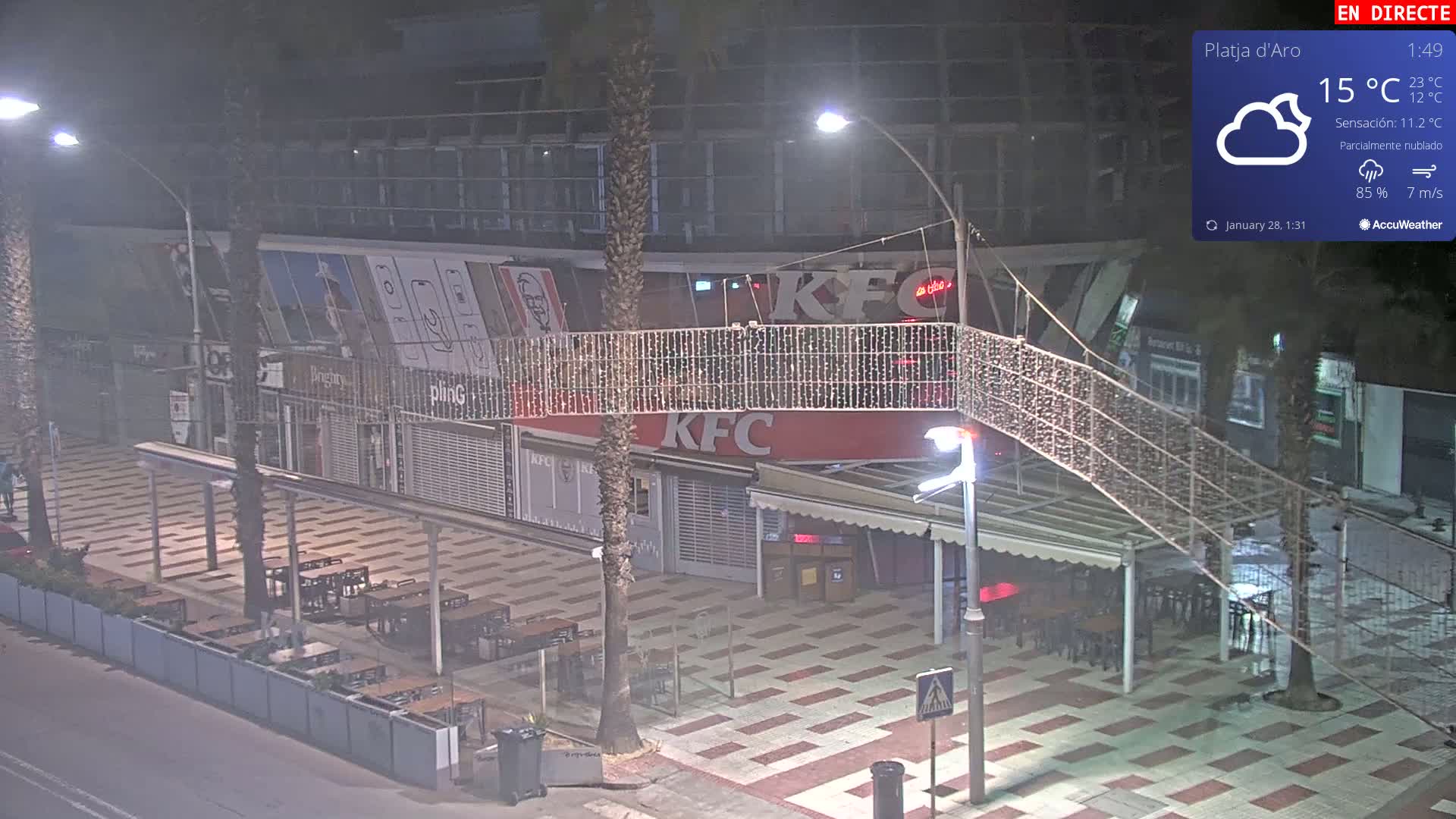 A nighttime, partly cloudy scene shows an outdoor plaza with several restaurants, including a KFC, and empty tables under string lights.