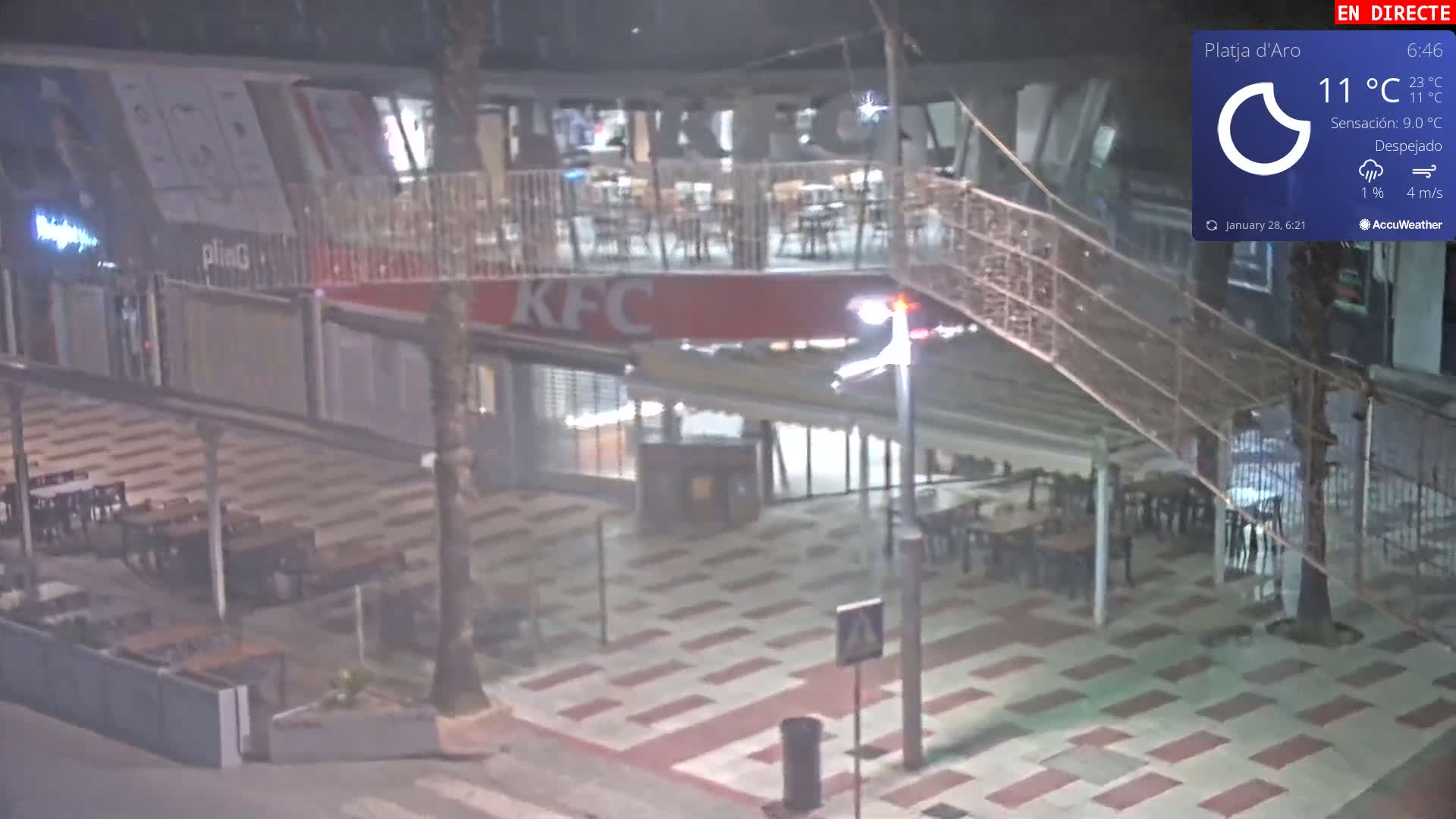 A nighttime, clear view of an outdoor seating area and a KFC restaurant, with a pedestrian bridge overhead.