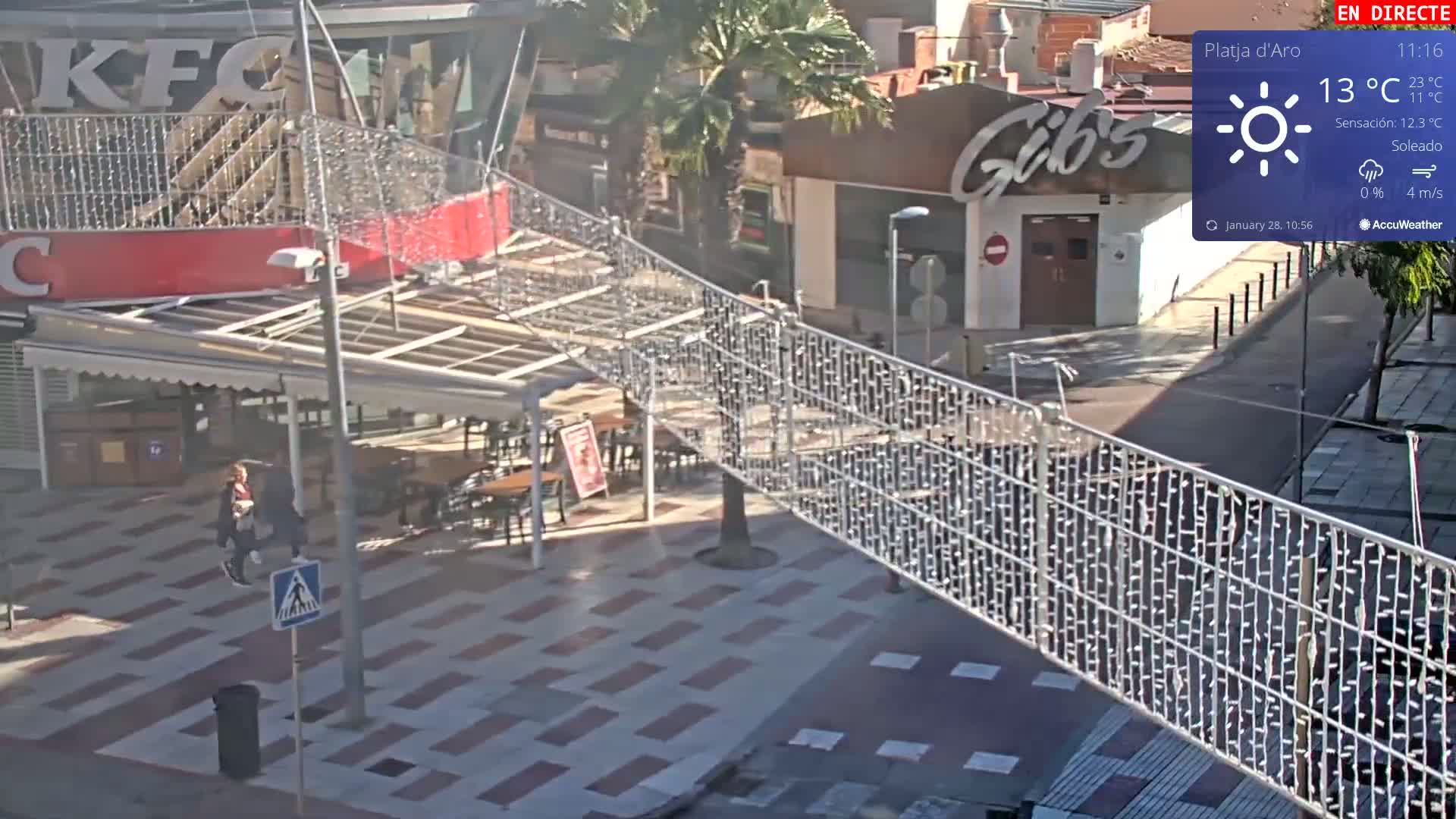 A sunny outdoor scene shows a pedestrian area with a metal fence strung with lights, a KFC, and a Gib's restaurant, with a few pedestrians visible.