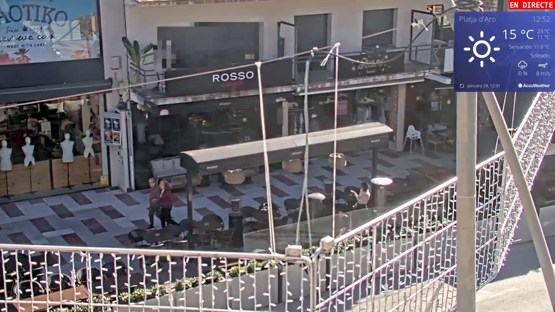 A sunny outdoor plaza with several shops and an outdoor cafe seating area, seen from an elevated viewpoint.