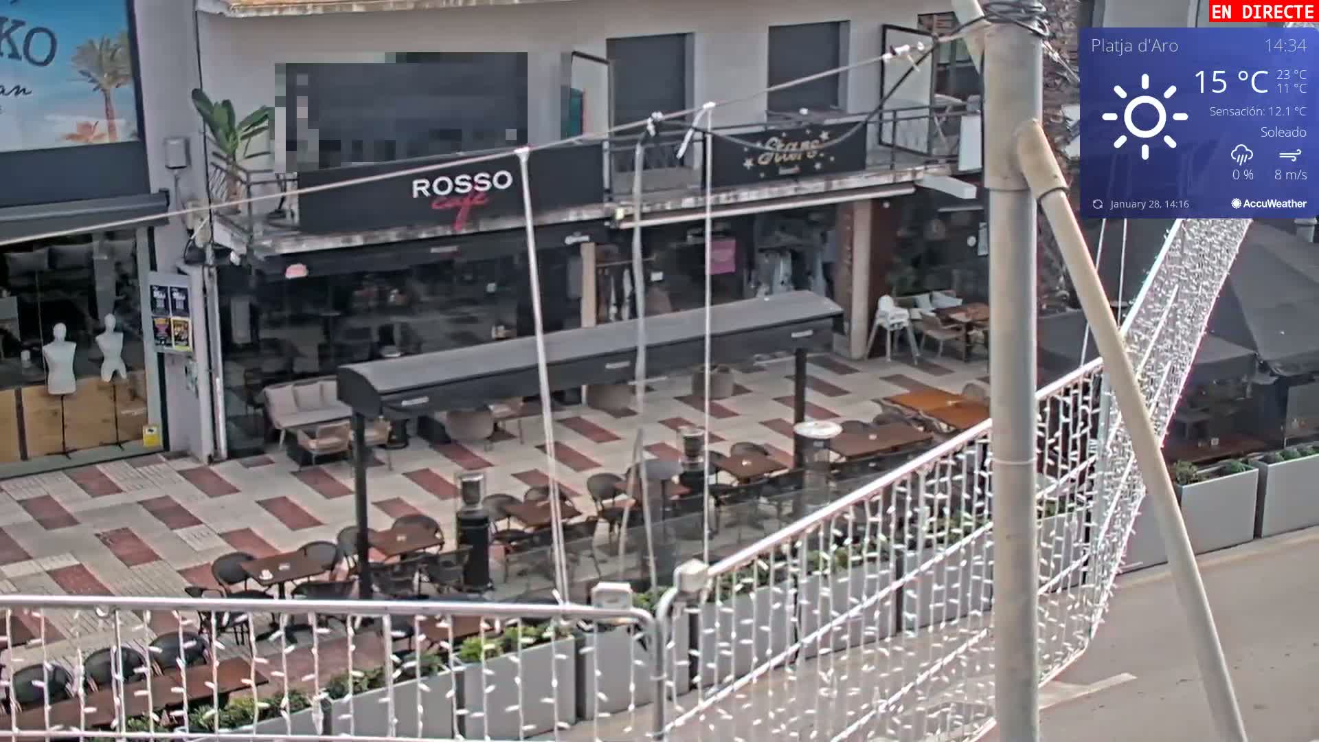A sunny outdoor cafe patio with several tables and chairs is seen from an elevated angle, with a street and additional buildings visible in the background.