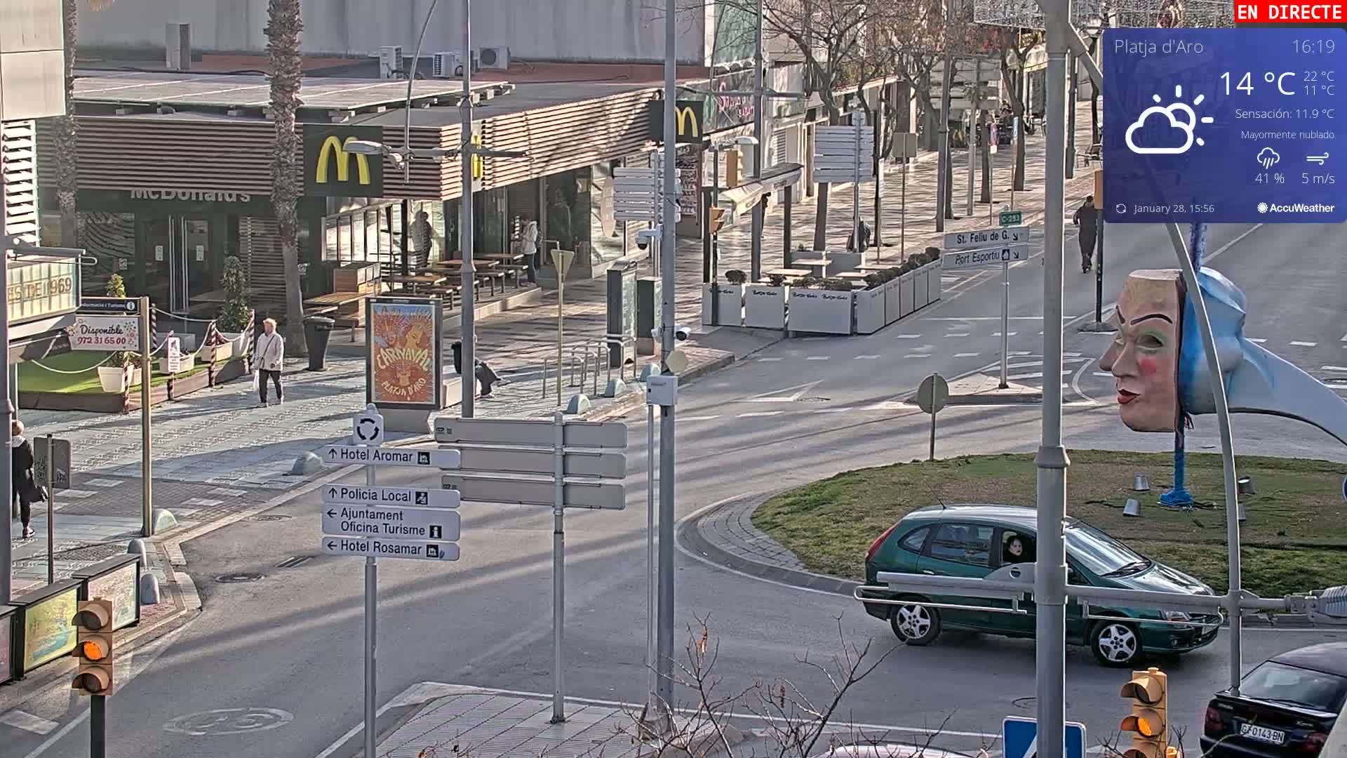 A partly cloudy daytime scene shows a street intersection with pedestrians, a green car, a McDonald's restaurant, and a whimsical sculpture of two faces.
