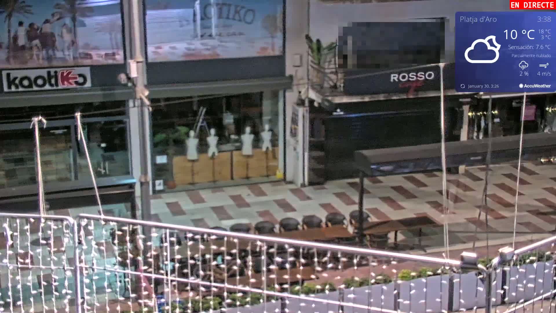 An outdoor plaza at night with partly cloudy skies, showing several shops with mannequins in the windows, and outdoor seating with a table and chairs.