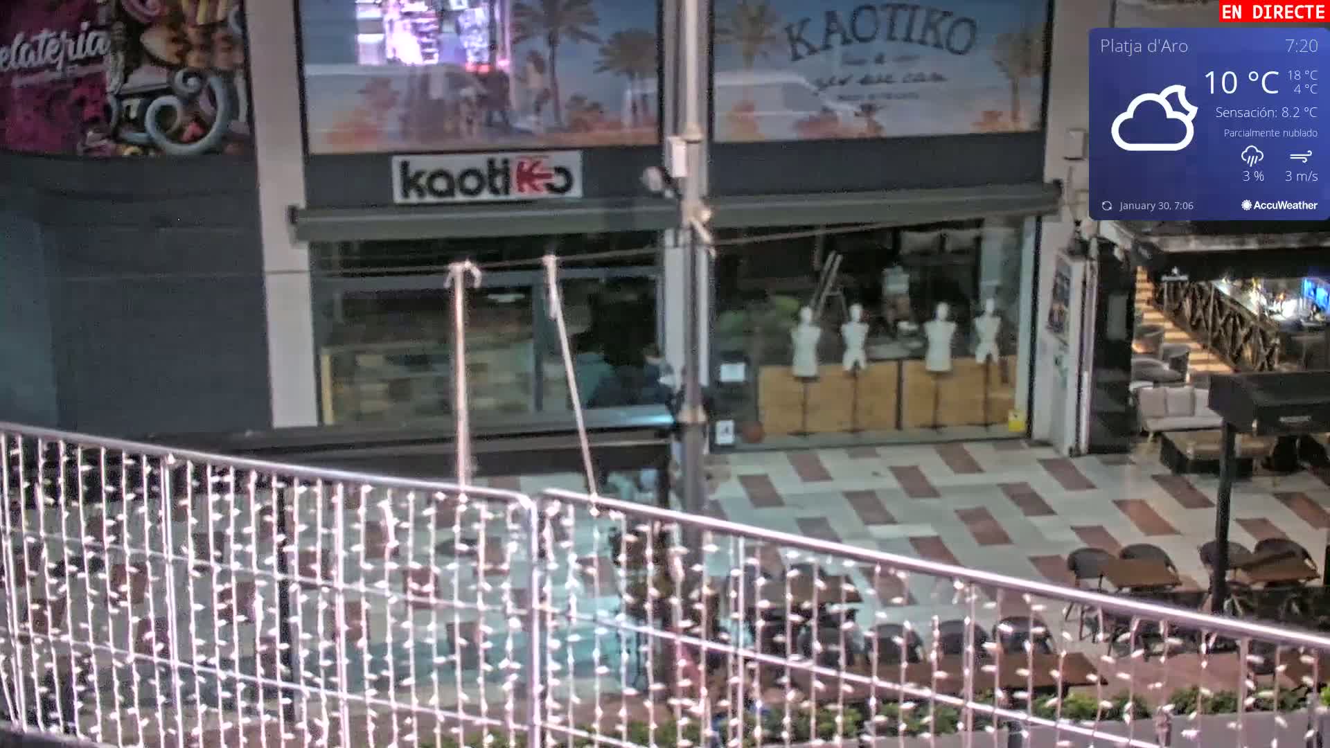A partially cloudy night scene shows an outdoor plaza with shops and restaurants, viewed from above a metal railing with small lights.