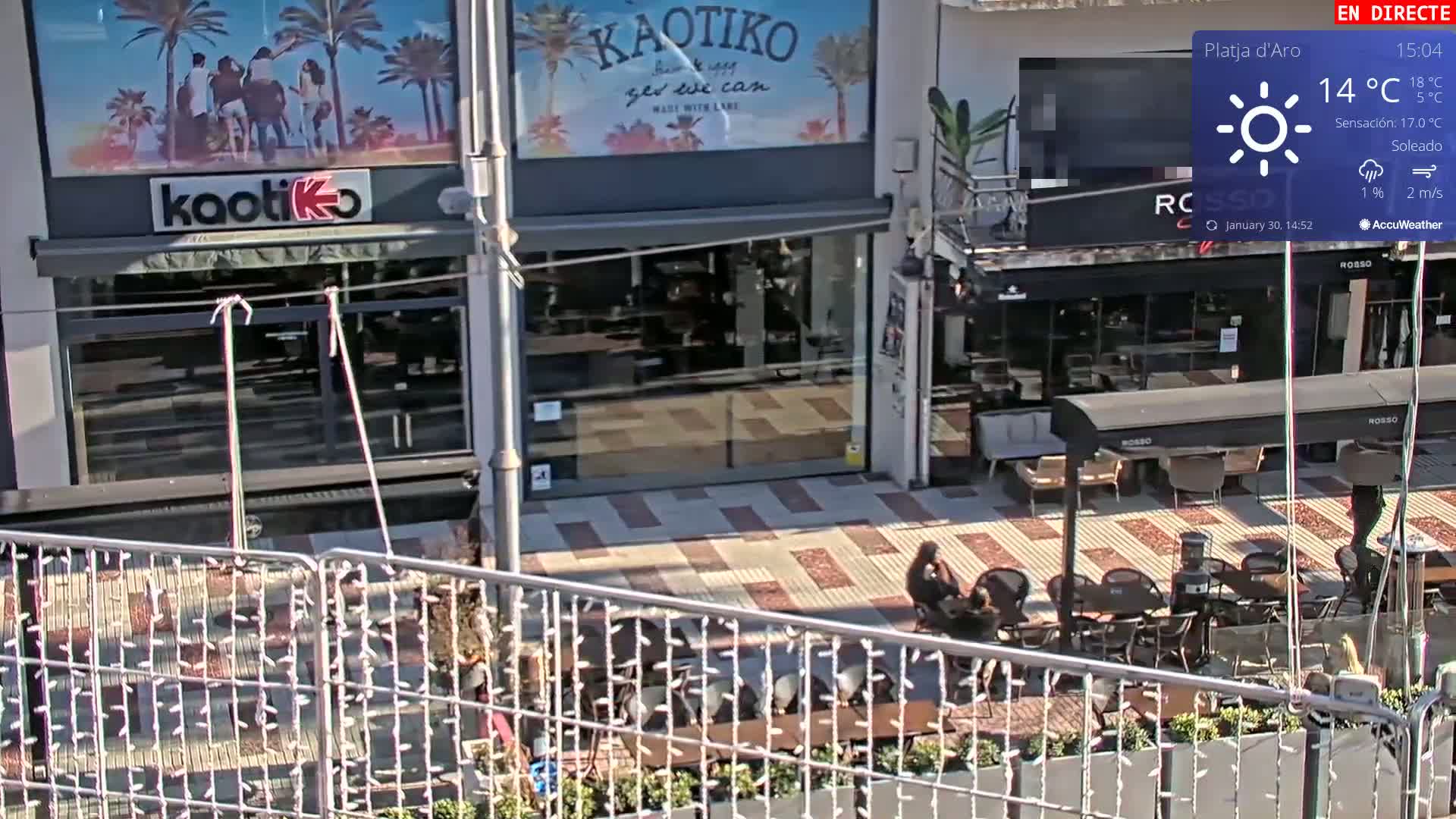 A sunny outdoor cafe scene with several occupied tables and chairs, situated in front of shops.