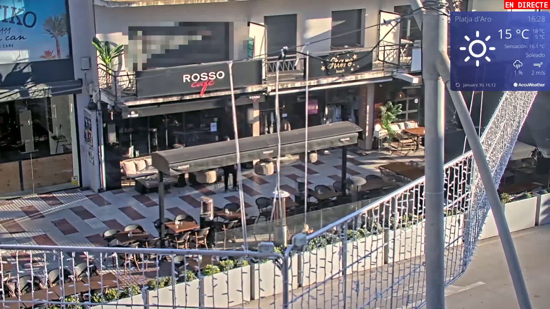 A sunny outdoor cafe patio with several tables and chairs, partially enclosed by a metal fence, is visible on a mostly sunny day.