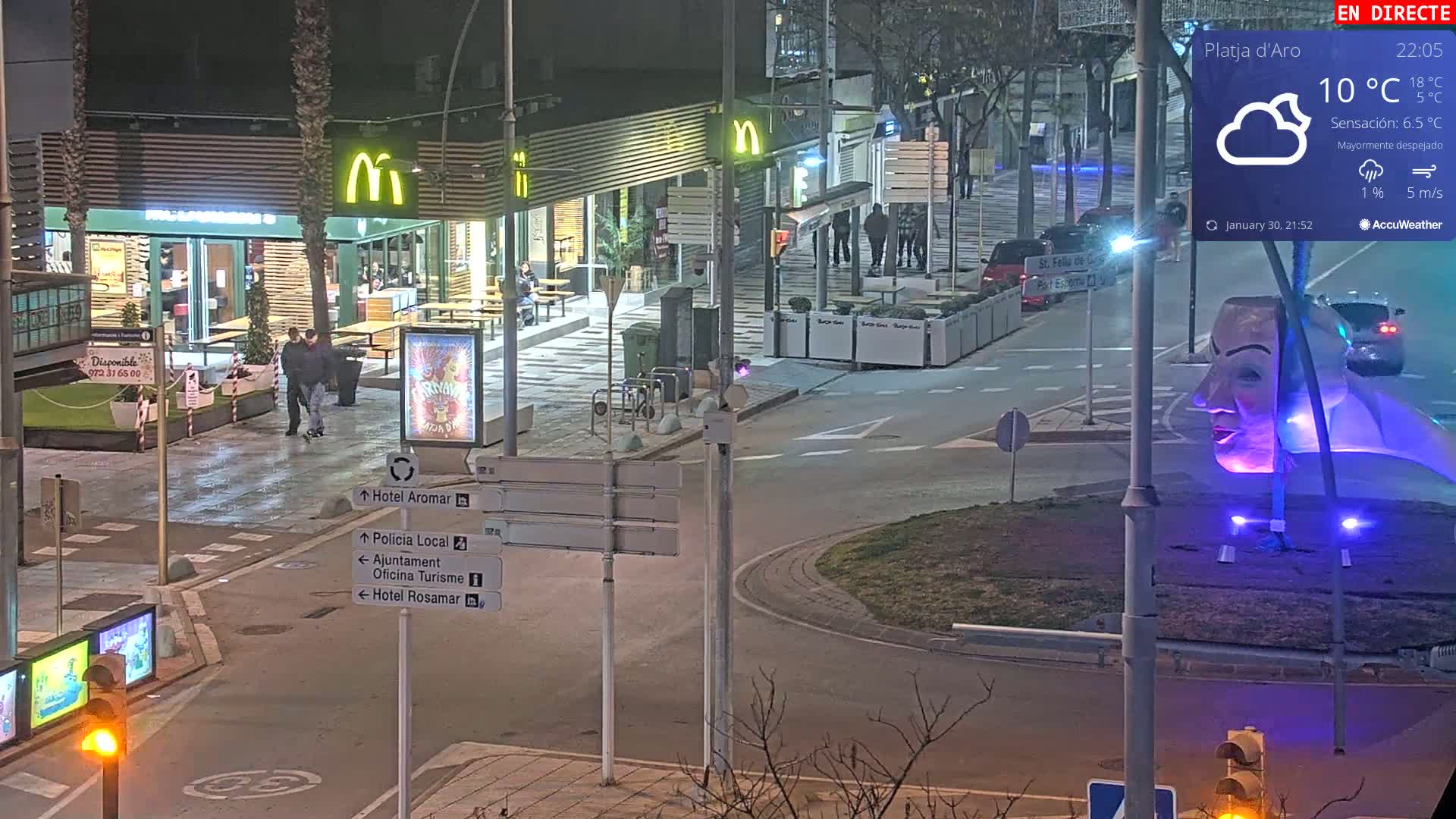 A nighttime street scene shows a McDonald's restaurant, a roundabout with a large illuminated sculpture of a face, and several pedestrians; the weather is mostly clear with a light breeze.