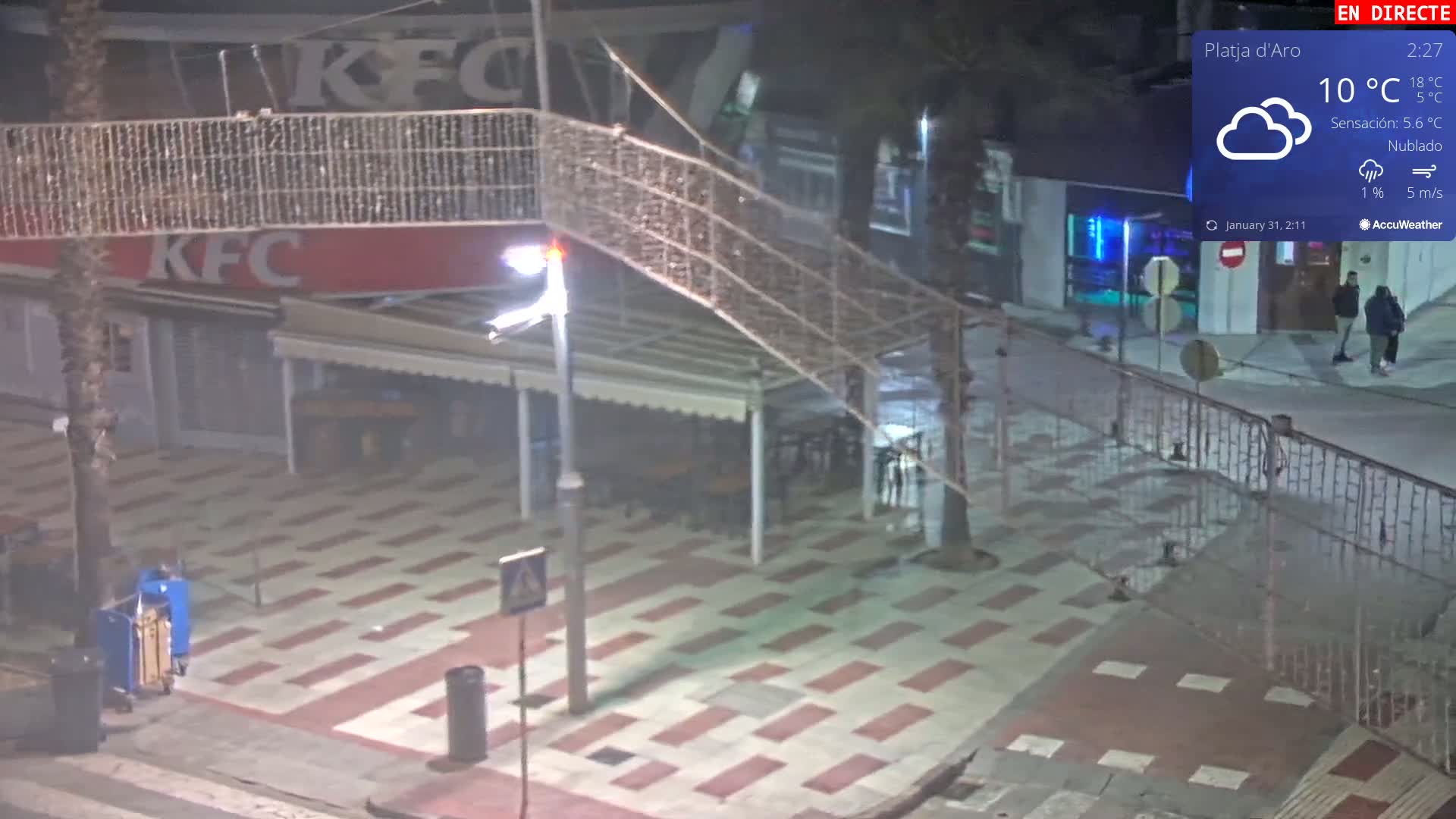 A mostly empty plaza at night under cloudy skies with a 10°C temperature, featuring a KFC restaurant and several people walking.