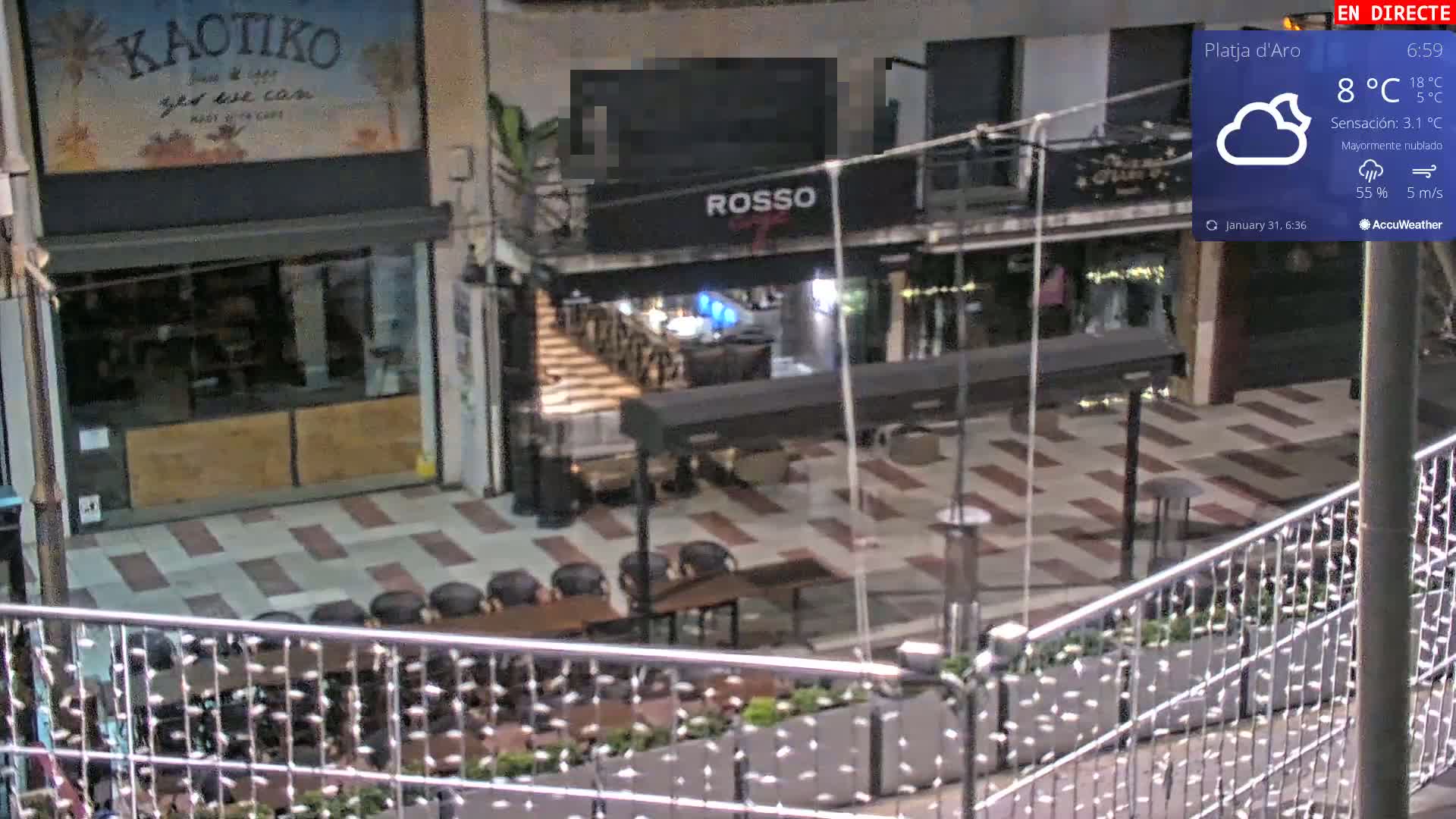 An outdoor plaza at night, mostly cloudy with a temperature of 8°C, features several shops and restaurants with empty tables and chairs.