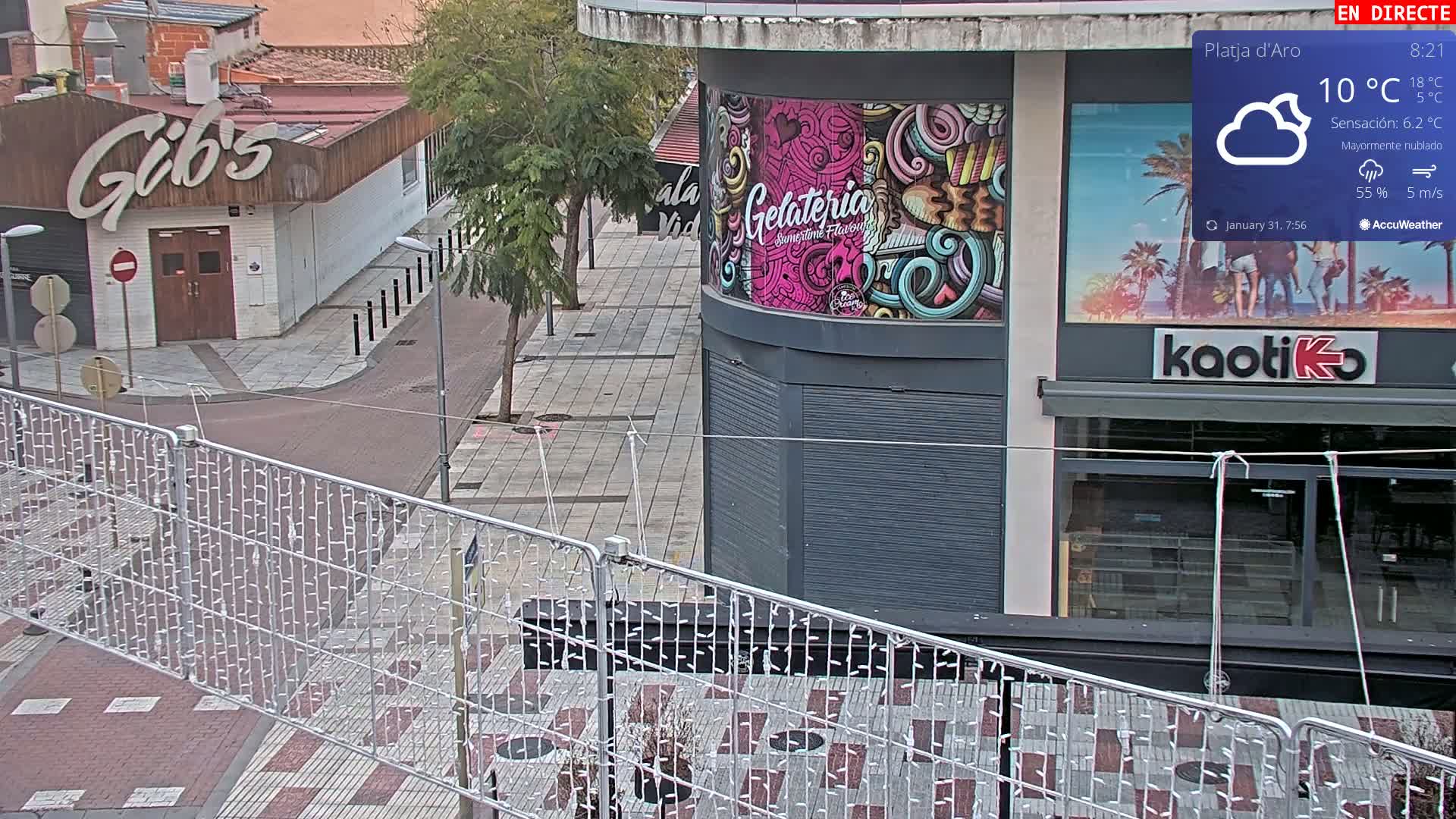 A mostly cloudy street scene shows a mostly empty plaza with a metal fence adorned with string lights in the foreground, and buildings with a gelato shop and bar visible in the background.