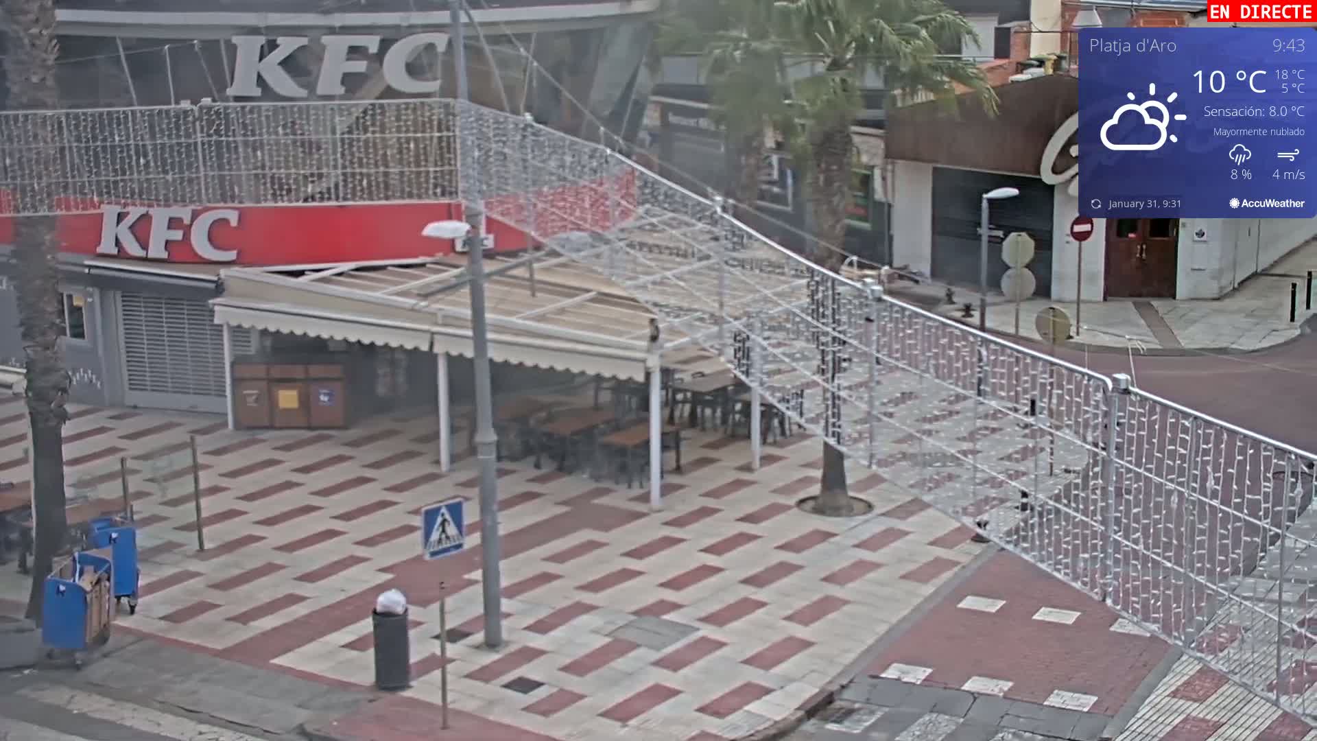 An outdoor scene on a mostly cloudy day shows a red-and-white KFC storefront with outdoor seating, partially enclosed by a metal fence decorated with white string lights.