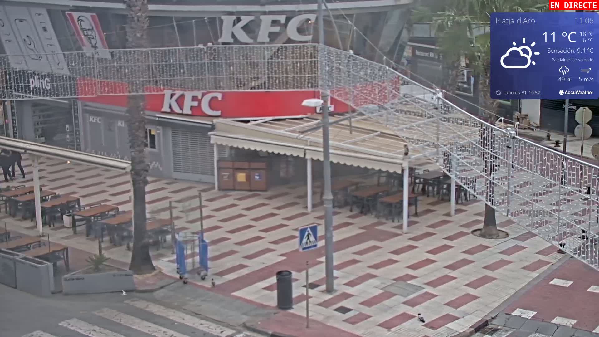 An outdoor paved area with tables and chairs outside a KFC restaurant under a partly sunny sky.