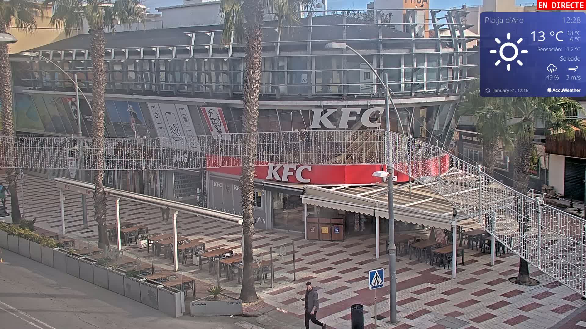 A sunny, outdoor scene shows a KFC restaurant with outdoor seating, a pedestrian walkway with string lights, and palm trees on a tiled plaza.