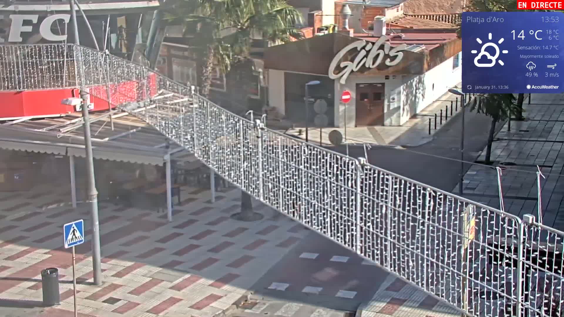 A mostly sunny street scene with a partially obstructed view of a street and buildings, featuring a metal fence decorated with string lights.