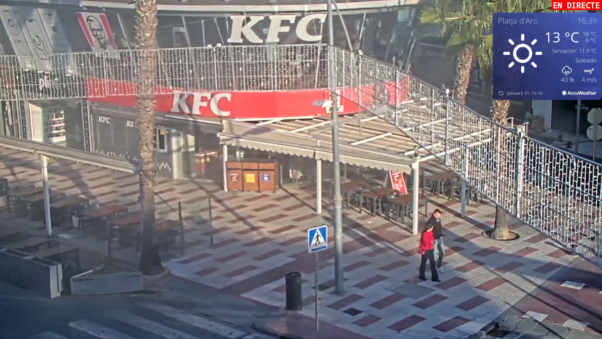A sunny, outdoor scene shows a paved area with tables and chairs outside a KFC, and two people walking by on a partly cloudy day with a 4 m/s wind.