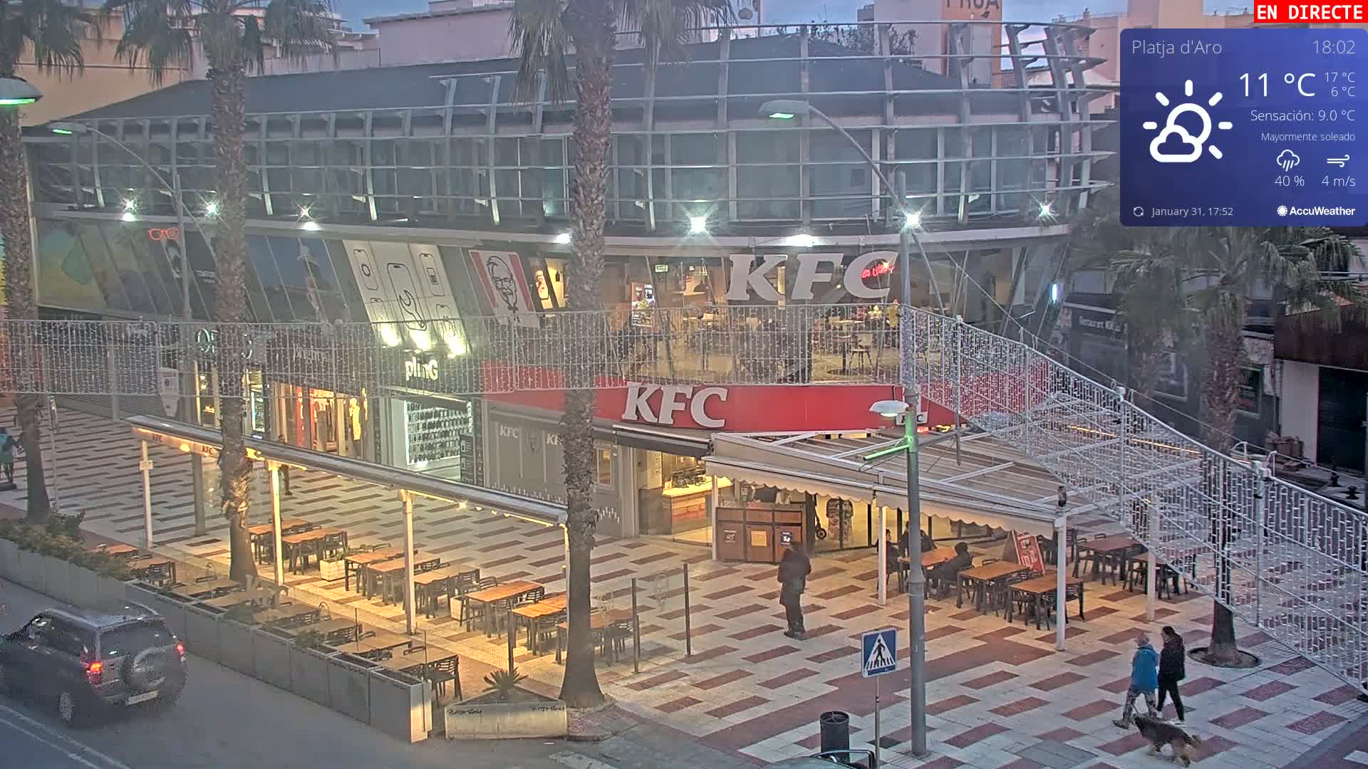 A mostly sunny evening scene shows a KFC restaurant with outdoor seating, a pedestrian walkway, and several people walking a dog.