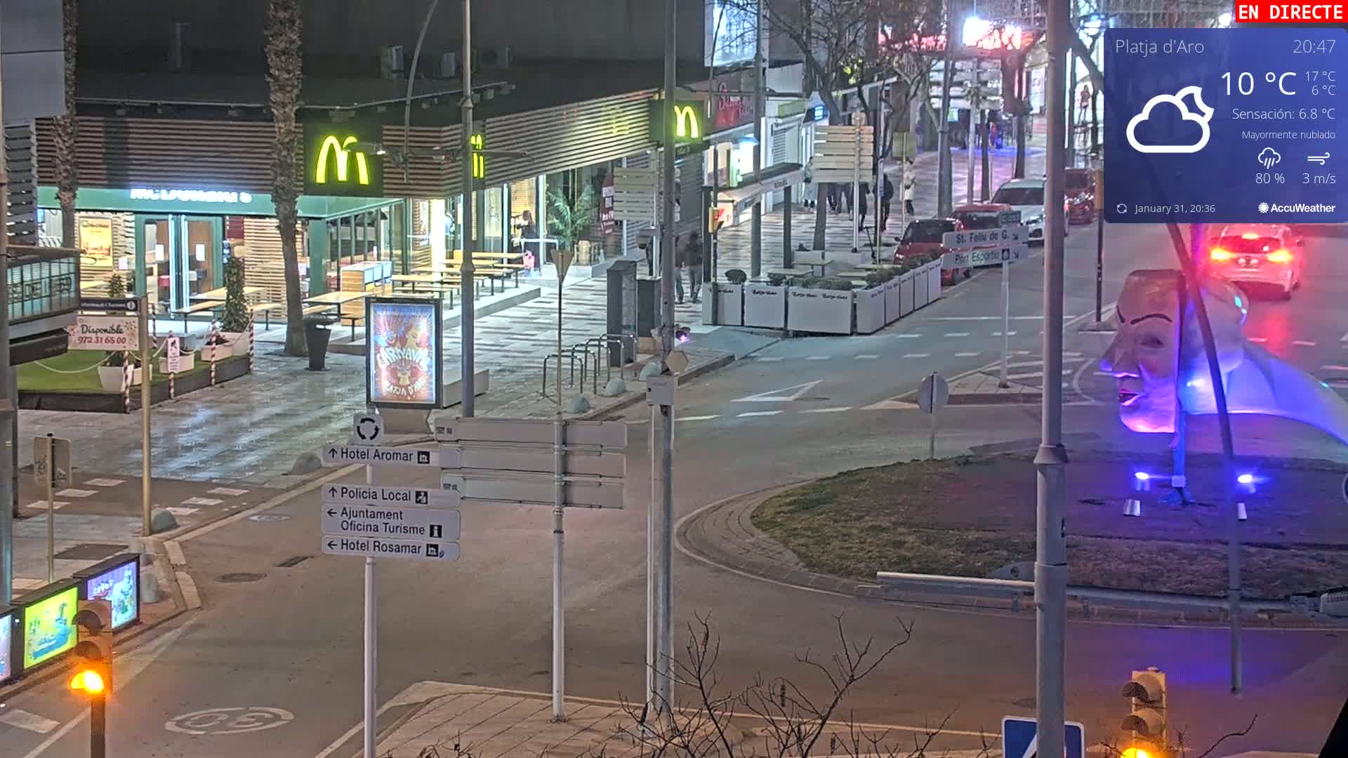 A nighttime street scene with mostly cloudy weather shows a McDonald's restaurant, outdoor seating, and a traffic circle with a large illuminated sculpture.